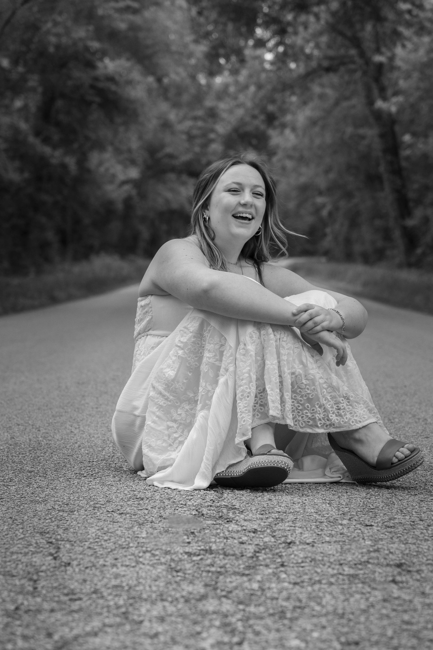A candid black and white portrait of someone sitting and laughing on a paved road surrounded by trees.