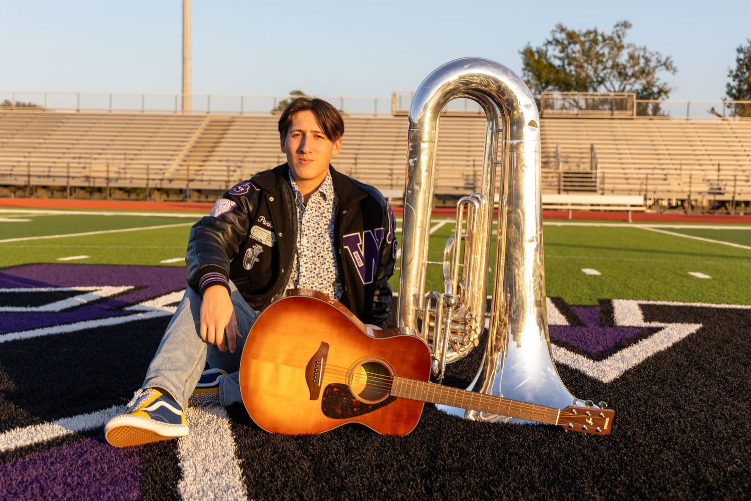 A young musician sits on a football field with an acoustic guitar and tuba in golden sunset light.