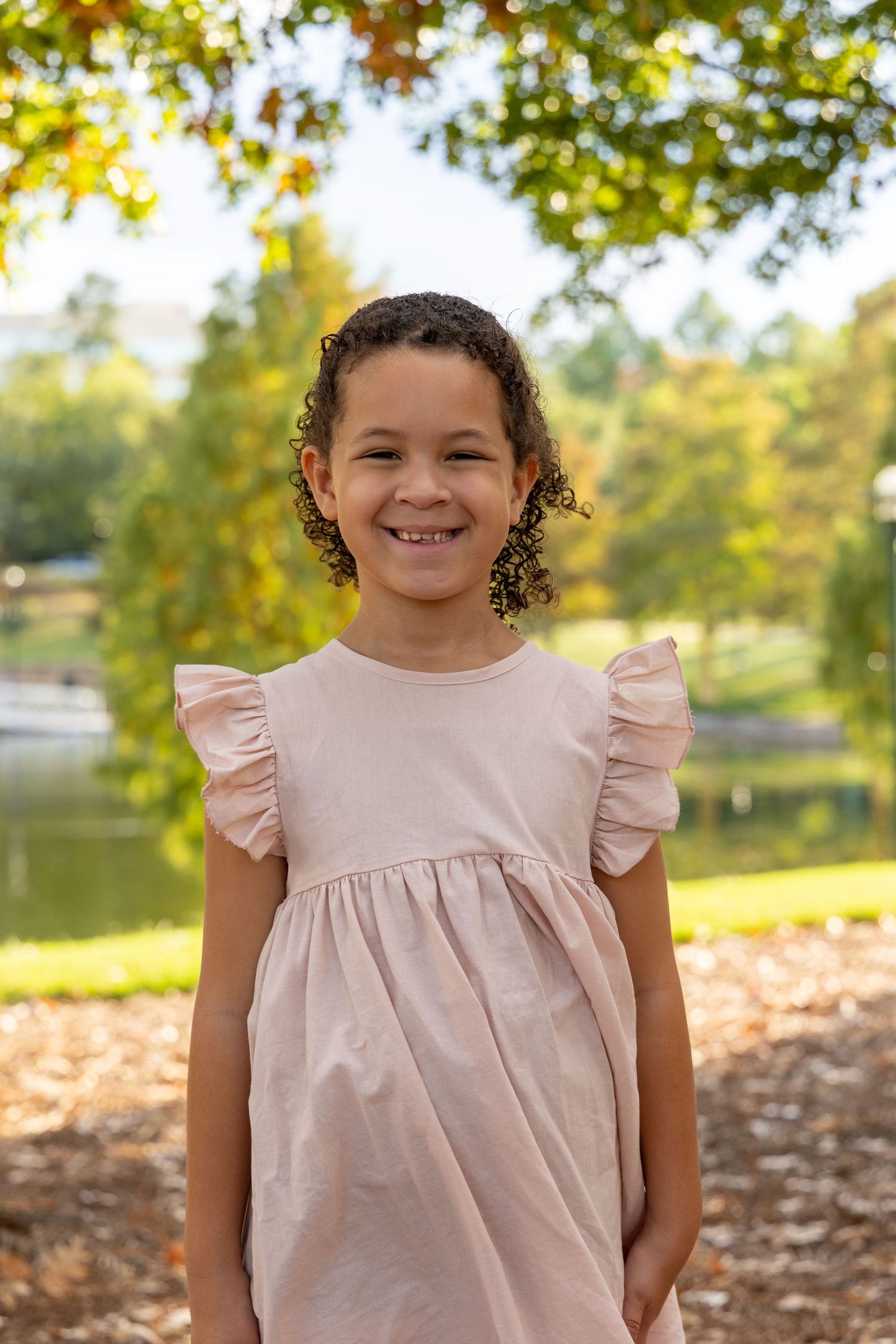 Smiling young girl in a pink ruffle dress standing outdoors in a park with autumn trees and a pond.