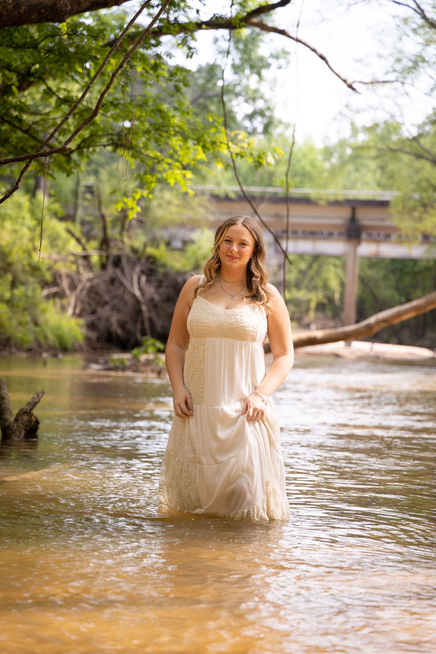 Young woman in white dress standing in shallow river with bridge and green trees in background.