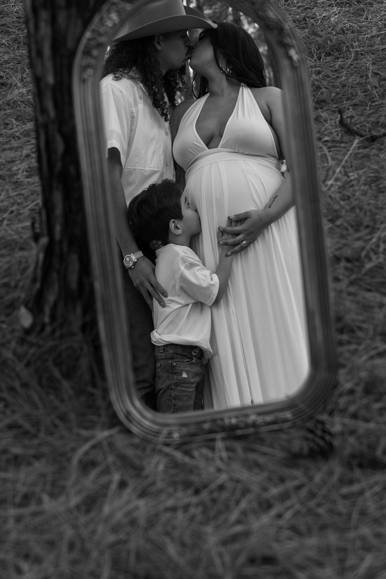 Black and white mirror reflection of pregnant woman, partner kissing, and child embracing belly outdoors.