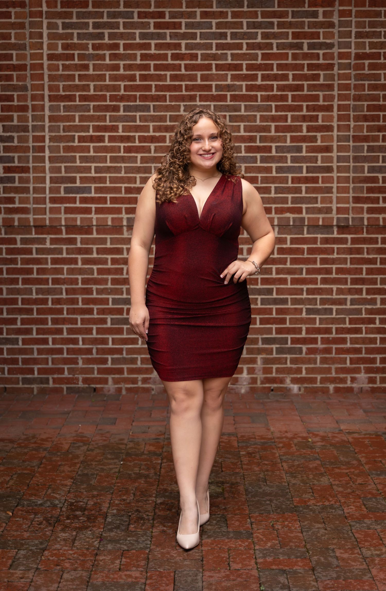 Young woman in a fitted burgundy dress posing confidently against a red brick wall on a cobblestone patio.