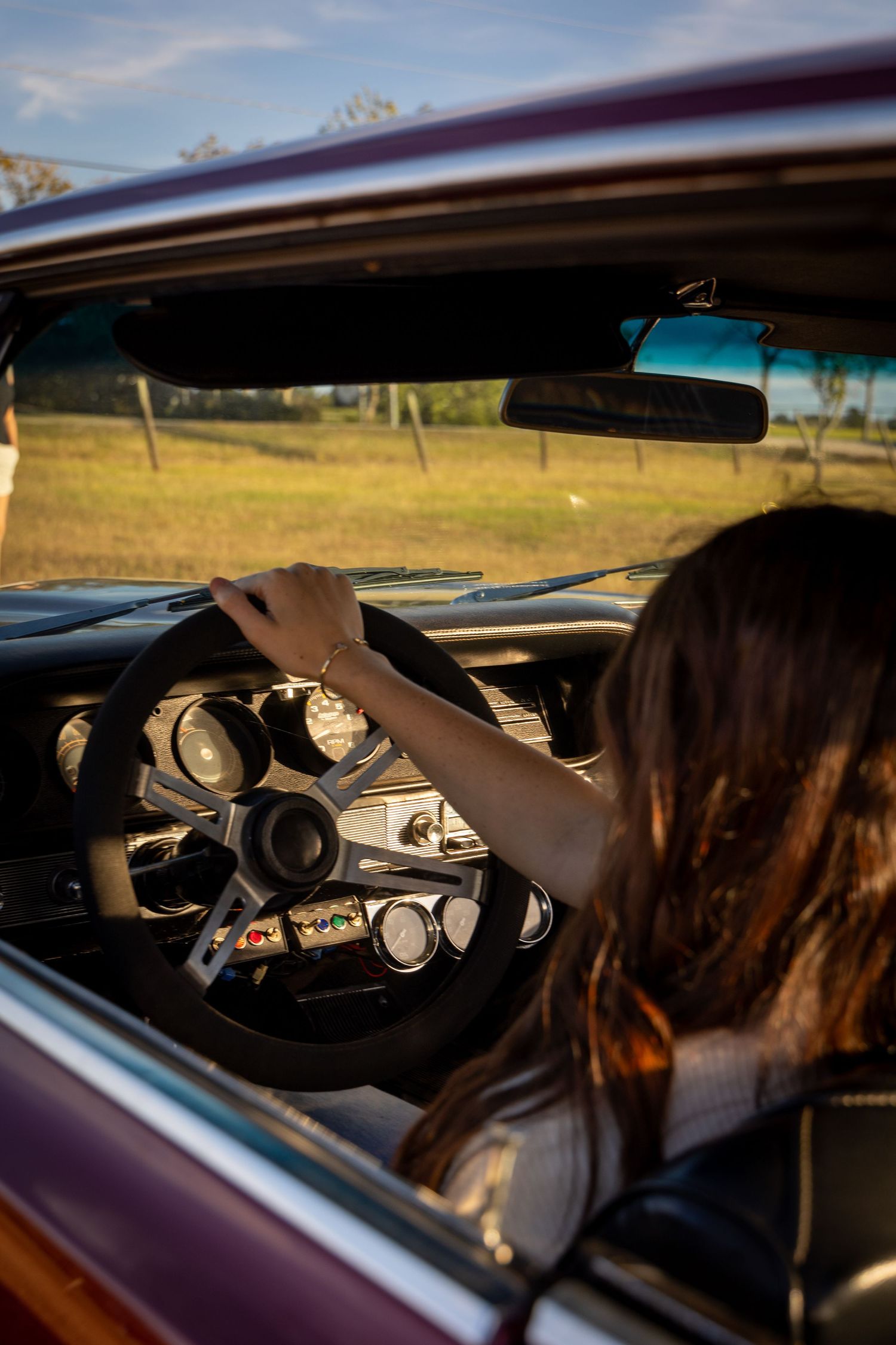 View through car window of someone driving a classic car with vintage dashboard gauges in warm sunlight.