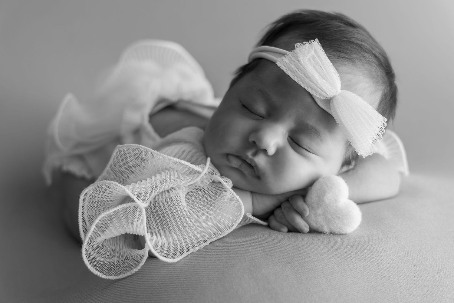 Black and white newborn photo of sleeping baby girl wearing white bow headband with ribbon decoration.
