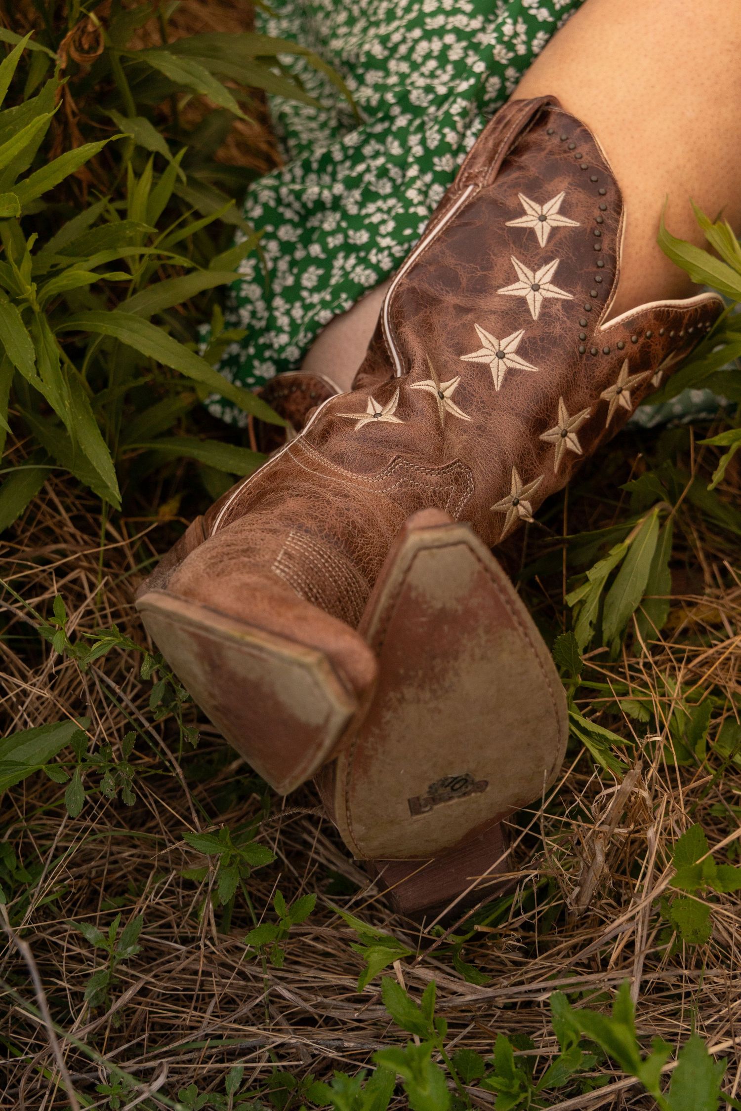 Brown leather cowboy boots with star embroidery, worn with a green floral dress, resting on grass and pine needles.
