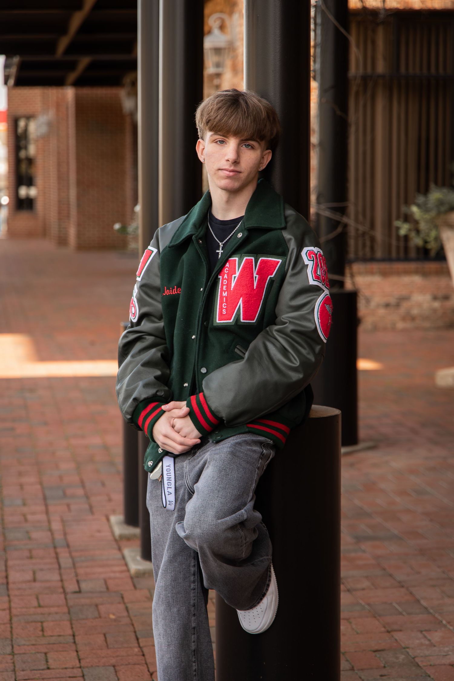 Young man in green varsity jacket with W letter patch leans against pole on brick sidewalk.