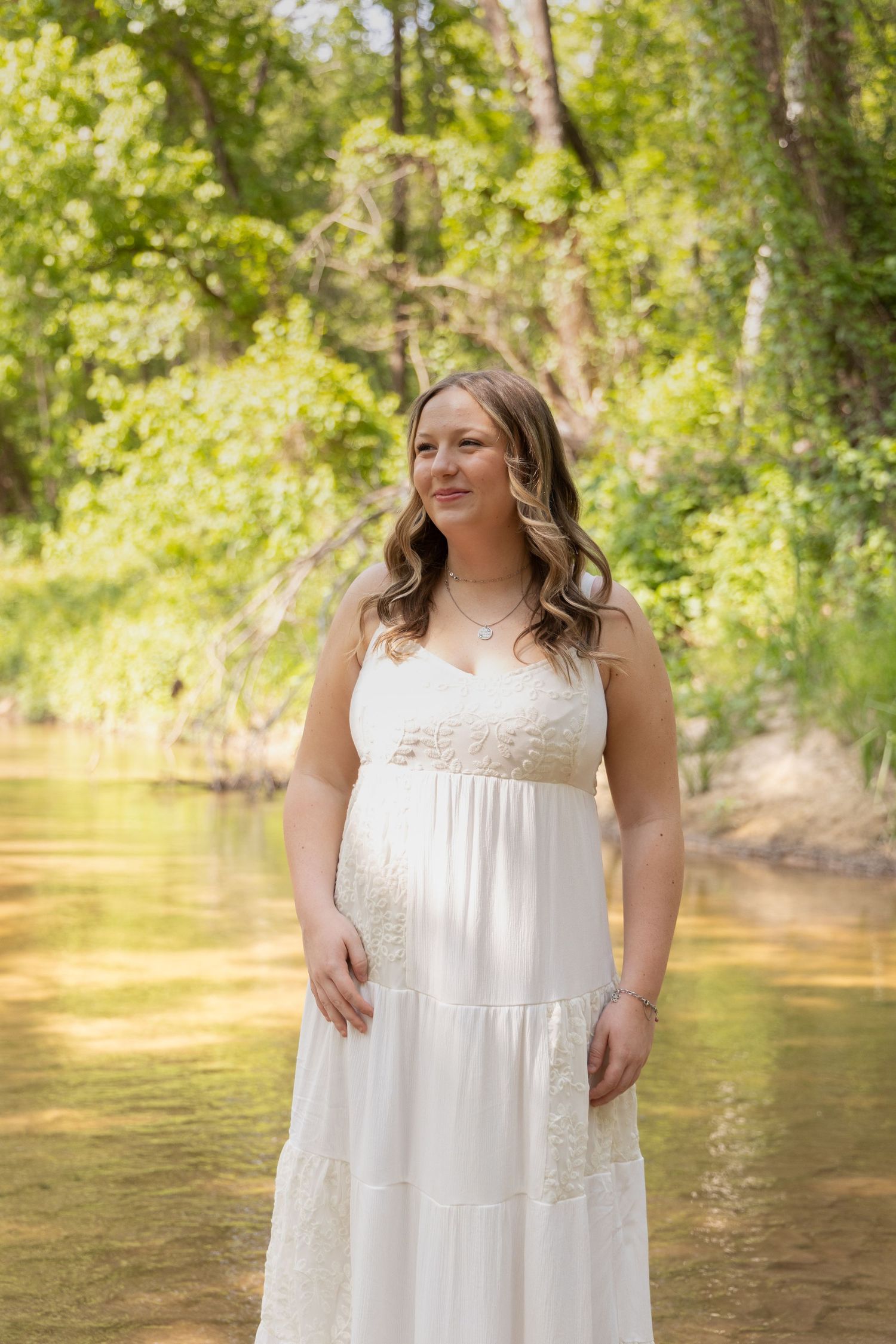 Pregnant woman in white lace sundress standing in a shallow creek surrounded by lush green trees.