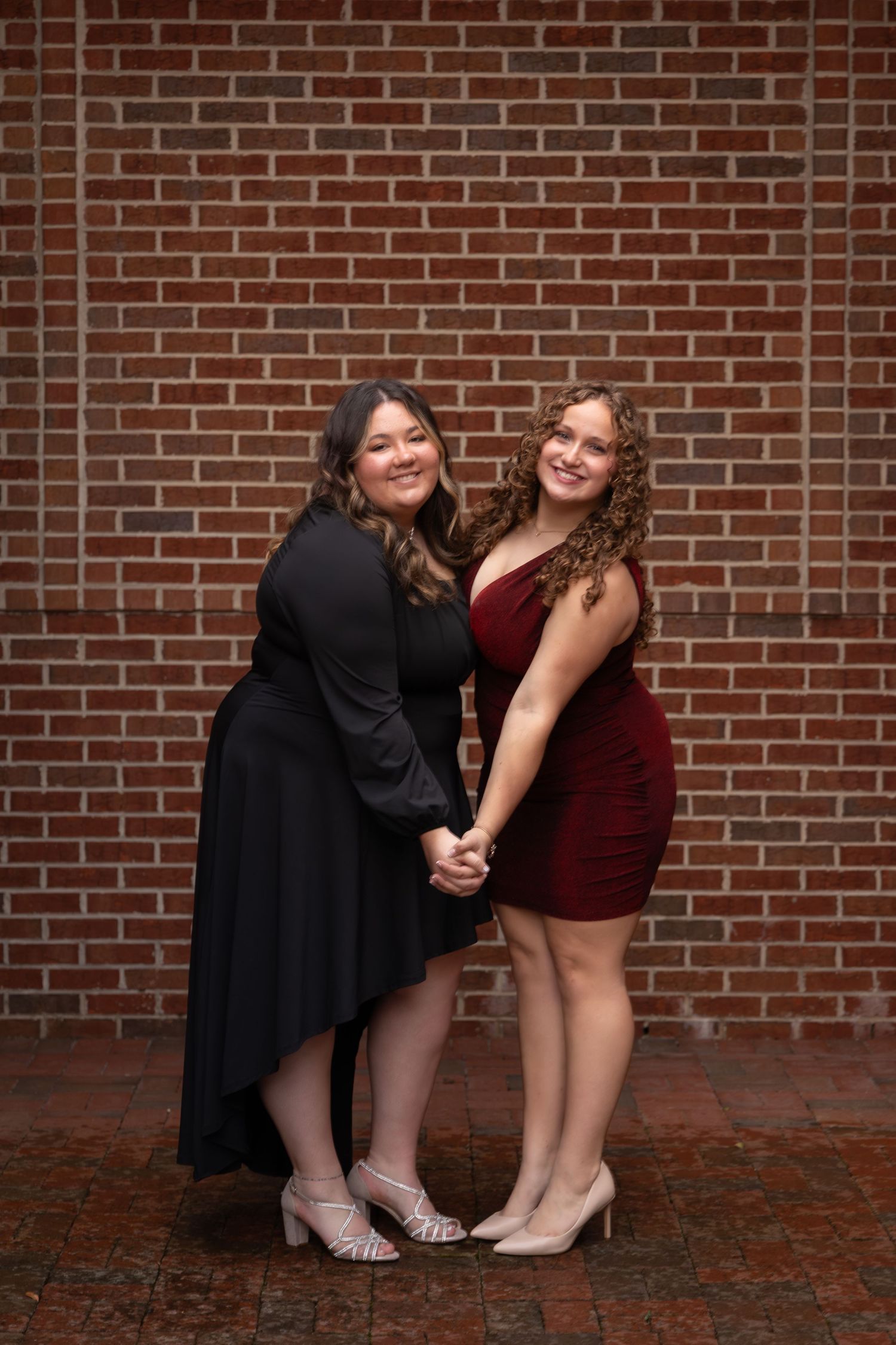 Two young women in formal dresses, one black and one red, hold hands and pose together in front of a brick wall.