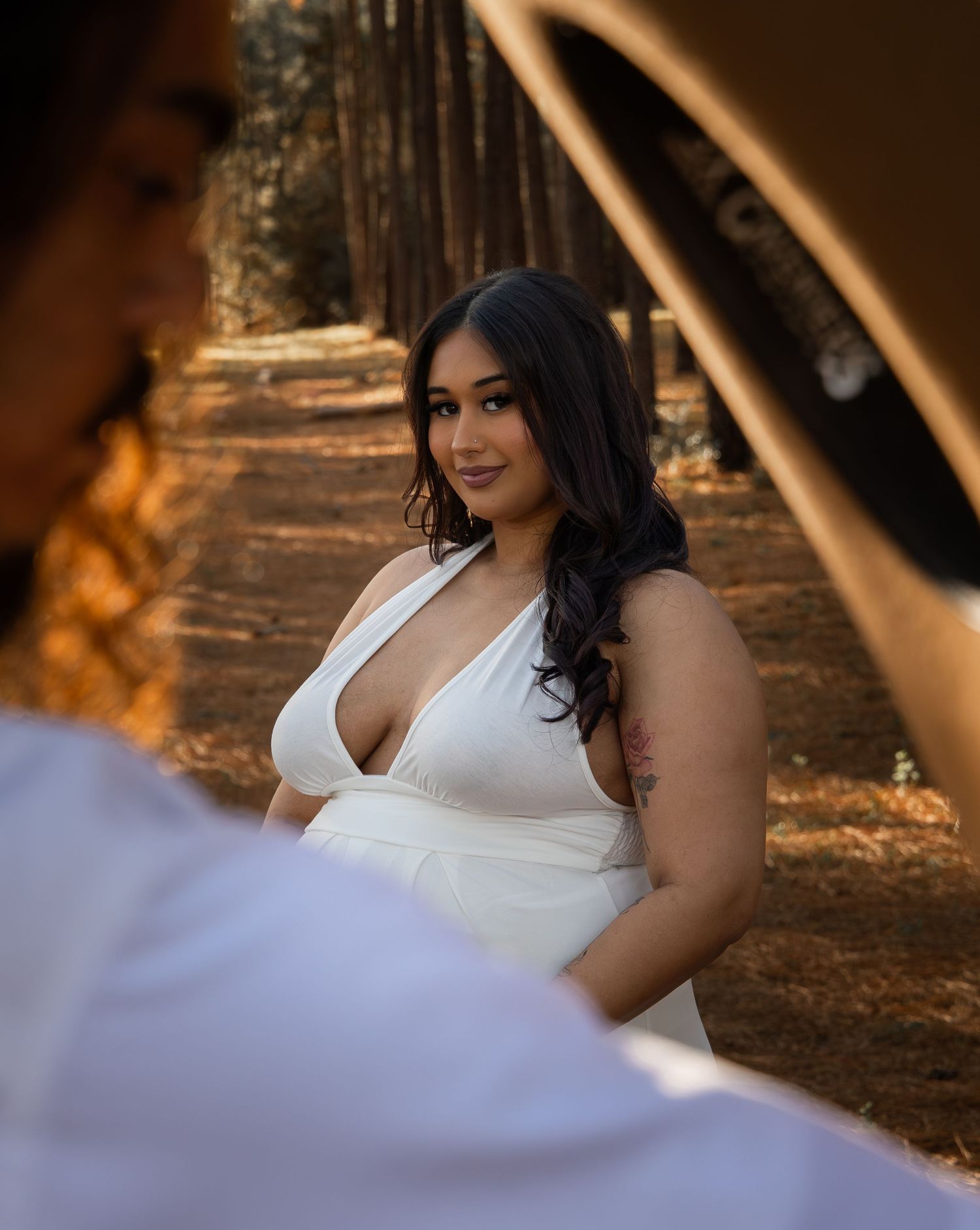 Woman in white deep-V dress poses in sunlit forest path, warm golden light filtering through tall pine trees.
