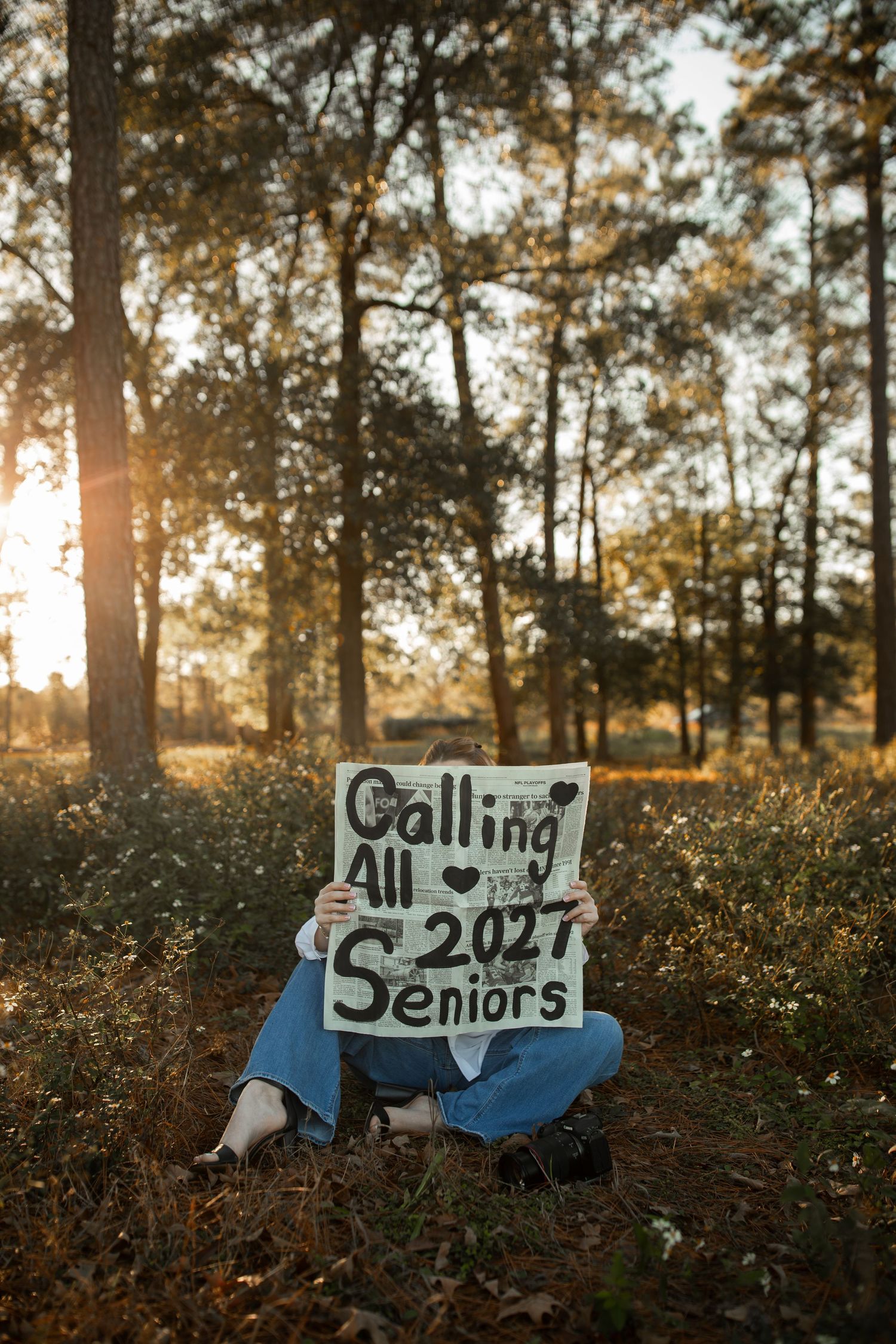 Person sitting outdoors holding a handmade sign reading Calling All 2027 Seniors in a sunlit forest setting.