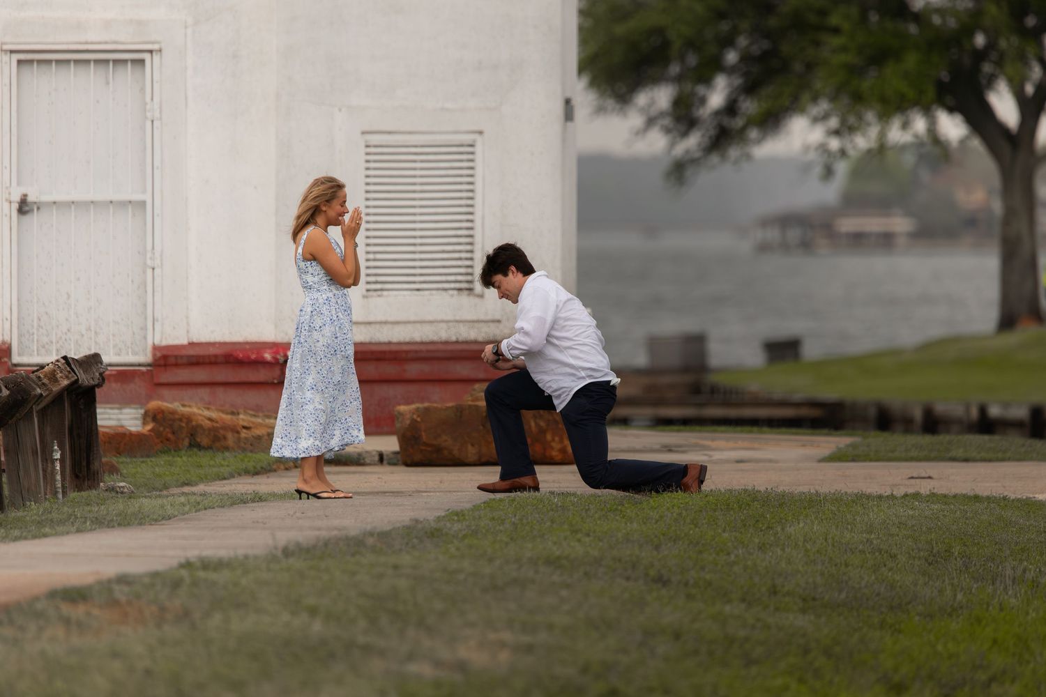 Man proposing on one knee to woman in blue dress outdoors near waterfront building.