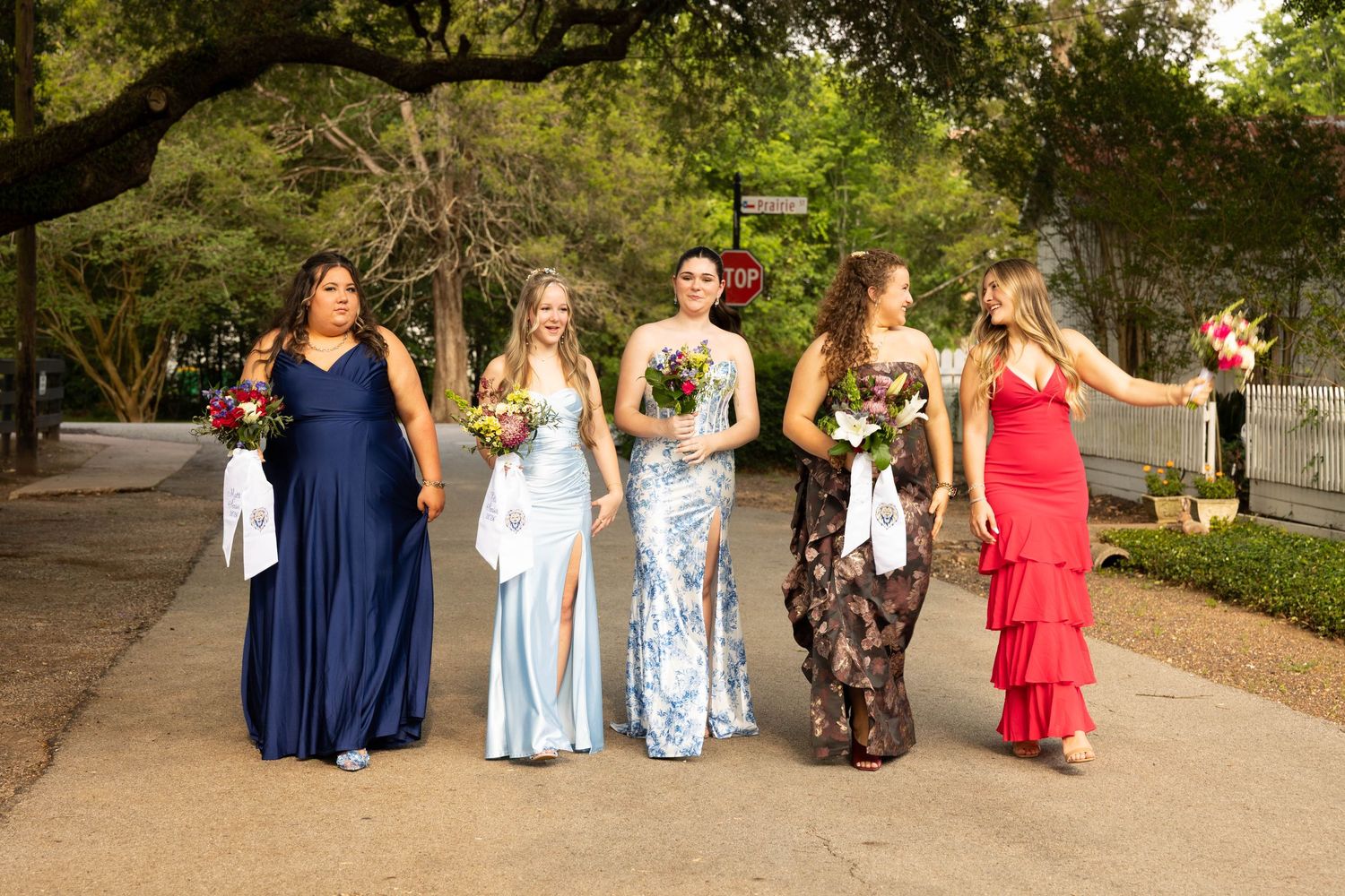 Five young women in formal gowns walking together outdoors holding bouquets.