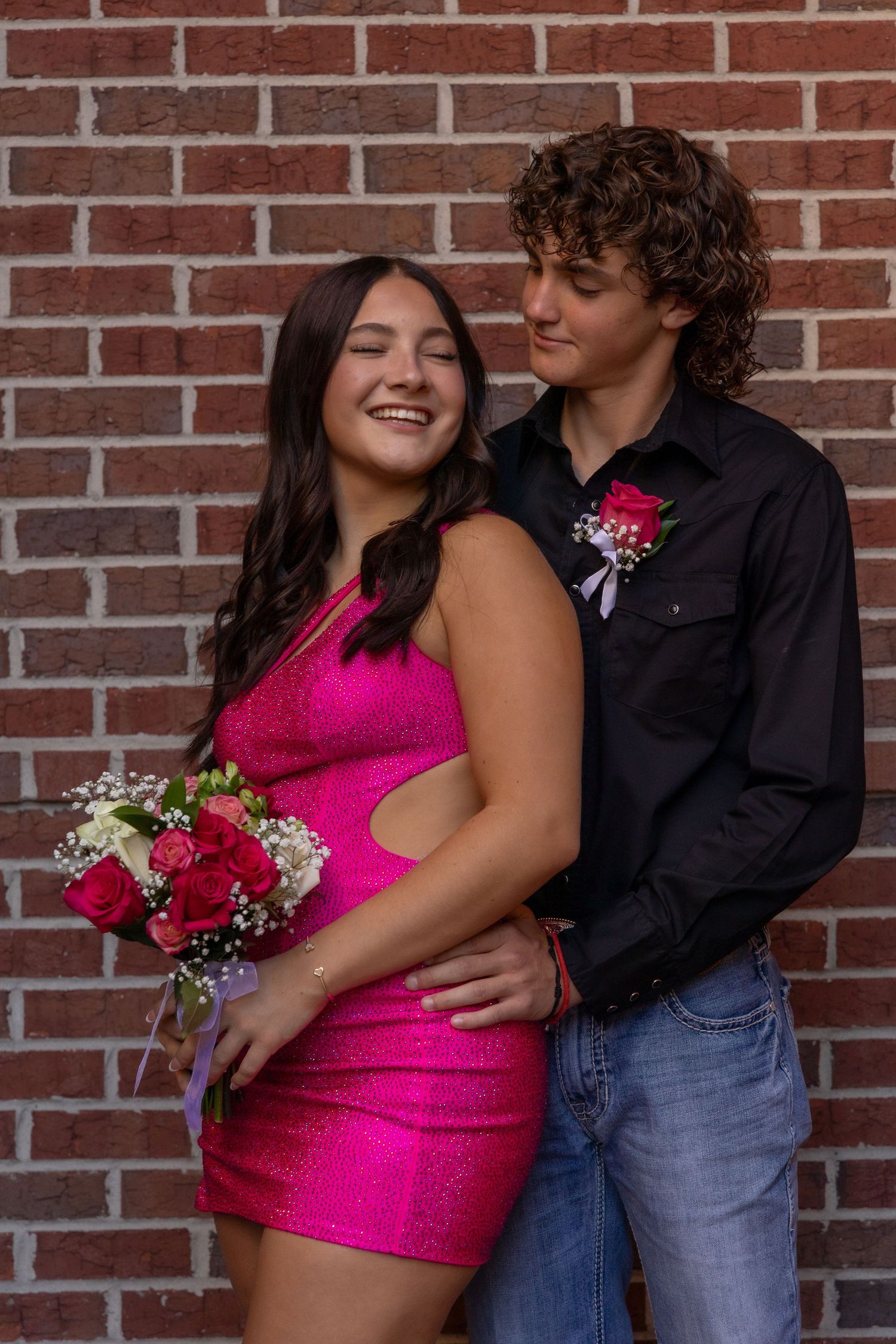 Teen couple in prom attire pose against brick wall, girl in pink dress holding bouquet, boy in black shirt with boutonniere.