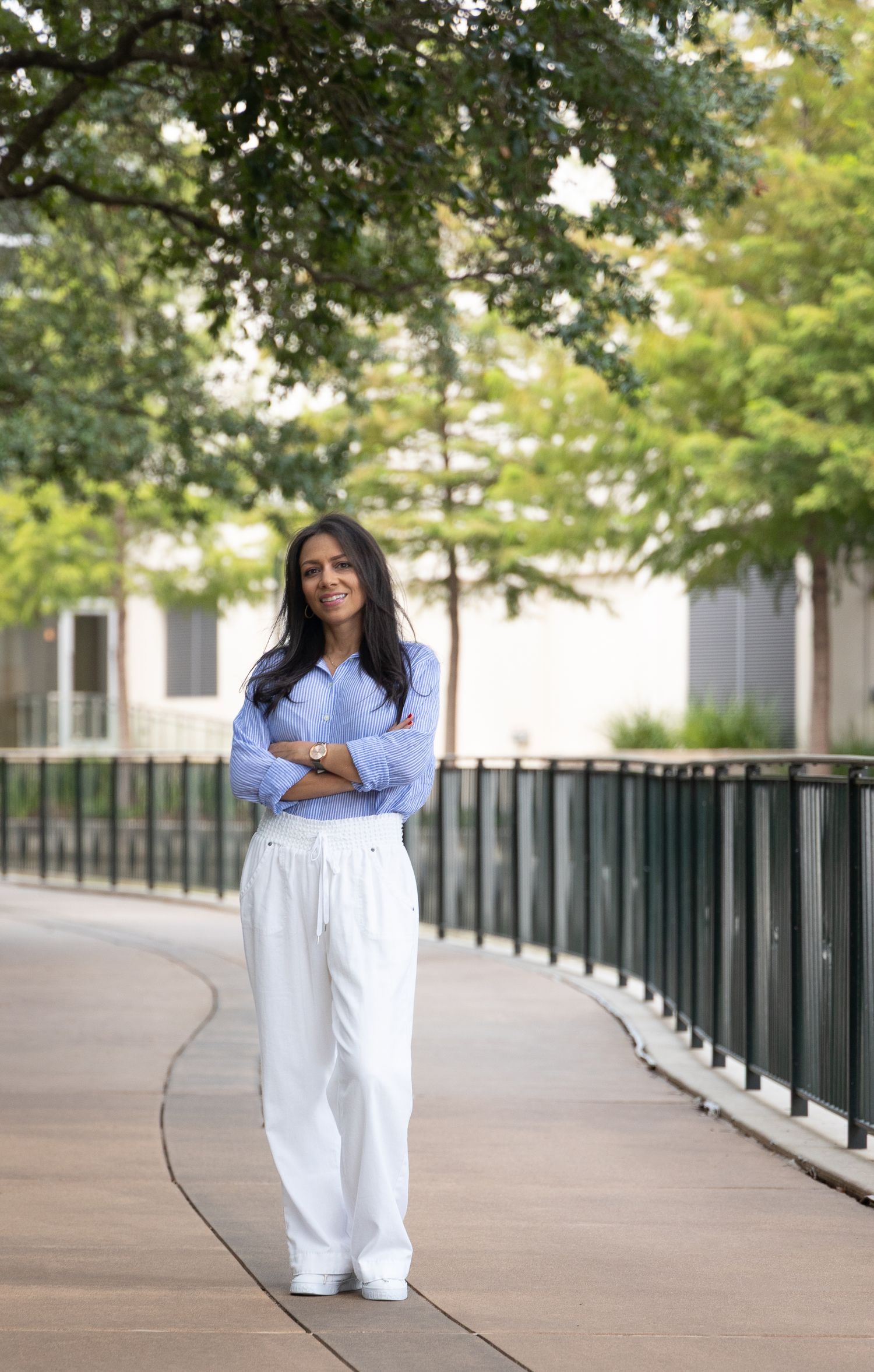 Woman in white trousers and blue shirt stands confidently on a walkway surrounded by lush green trees.