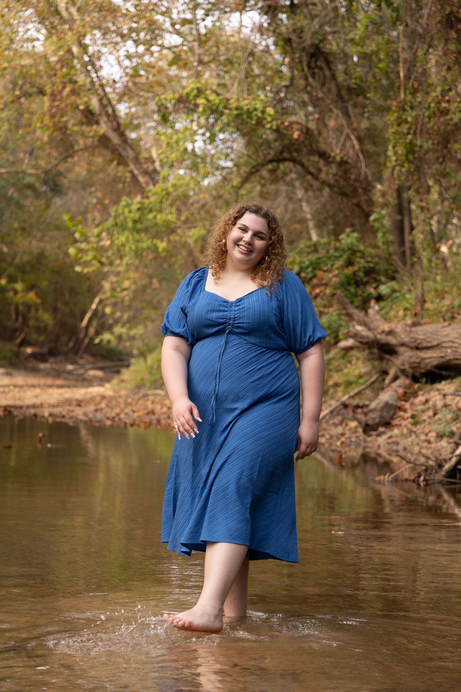 A smiling plus-size woman in a blue midi dress wades barefoot in a shallow forest creek.