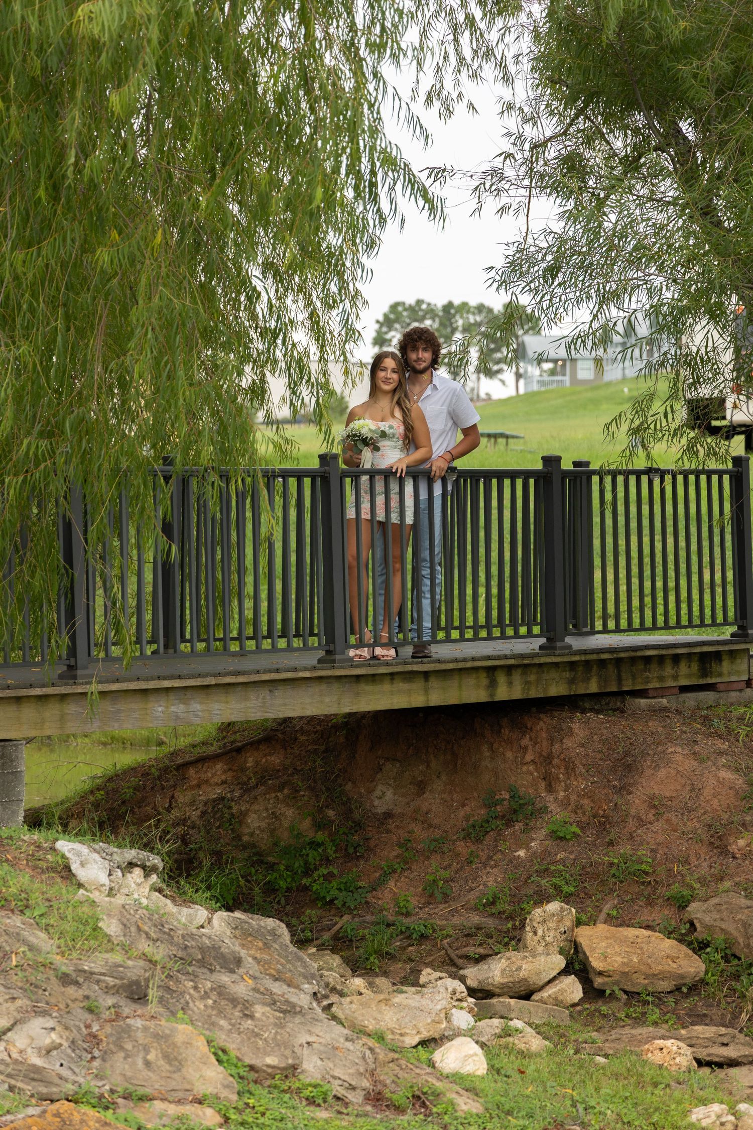 Couple embracing on a wooden bridge surrounded by lush green trees and rocky creek bed in a park setting.