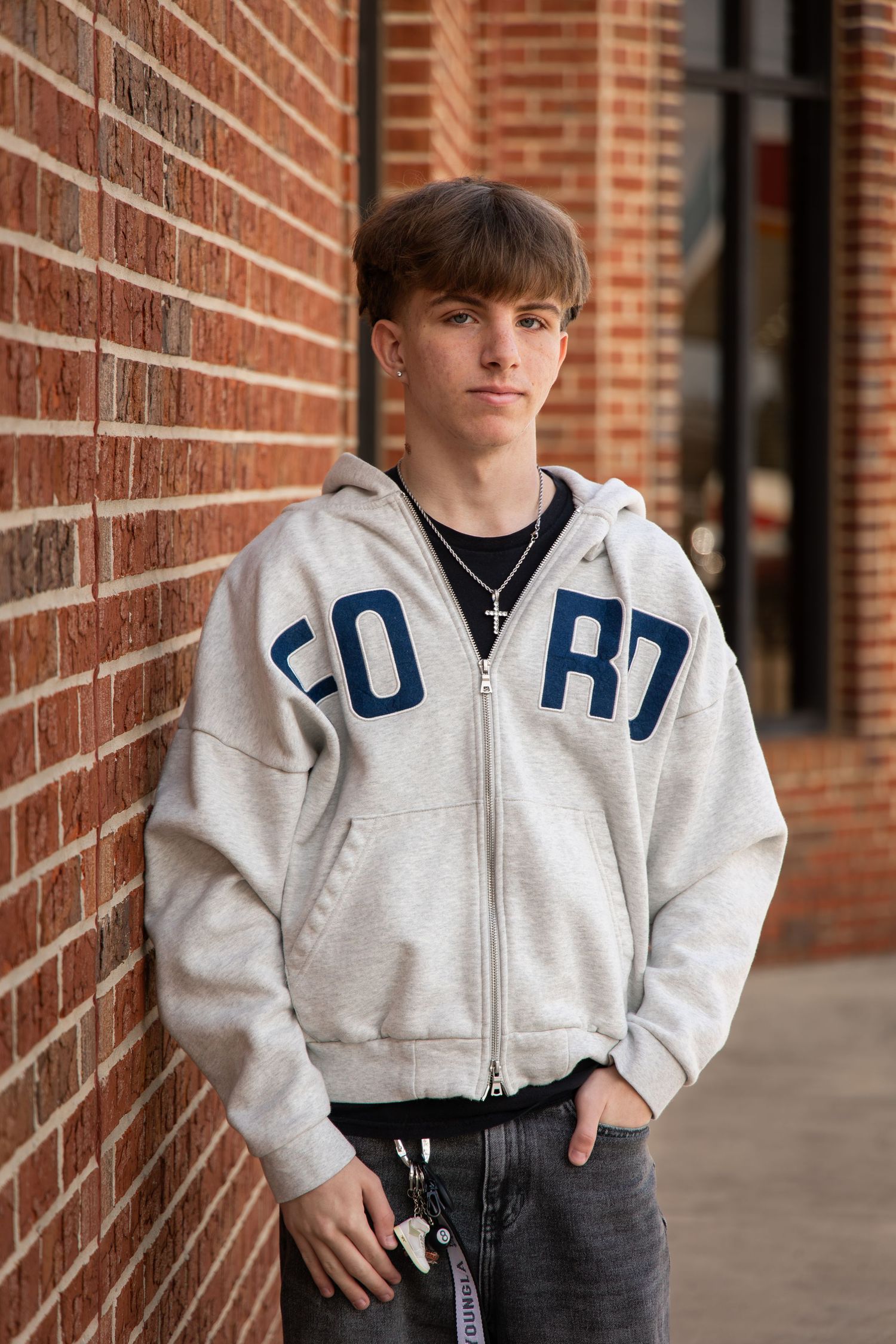 Teen boy in gray zip-up hoodie with blue letters leans against red brick wall outdoors.