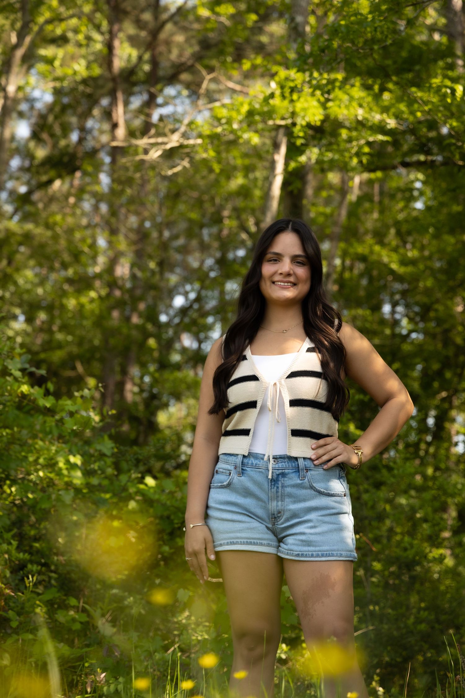 Young woman in striped crop top and denim shorts smiling outdoors in a lush green forest setting.