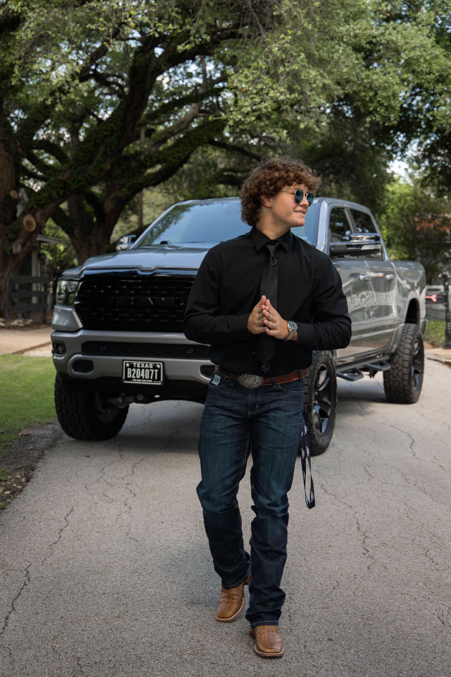 Young man in black shirt, jeans, and boots poses in front of a gray Ram pickup truck on a tree-lined street.