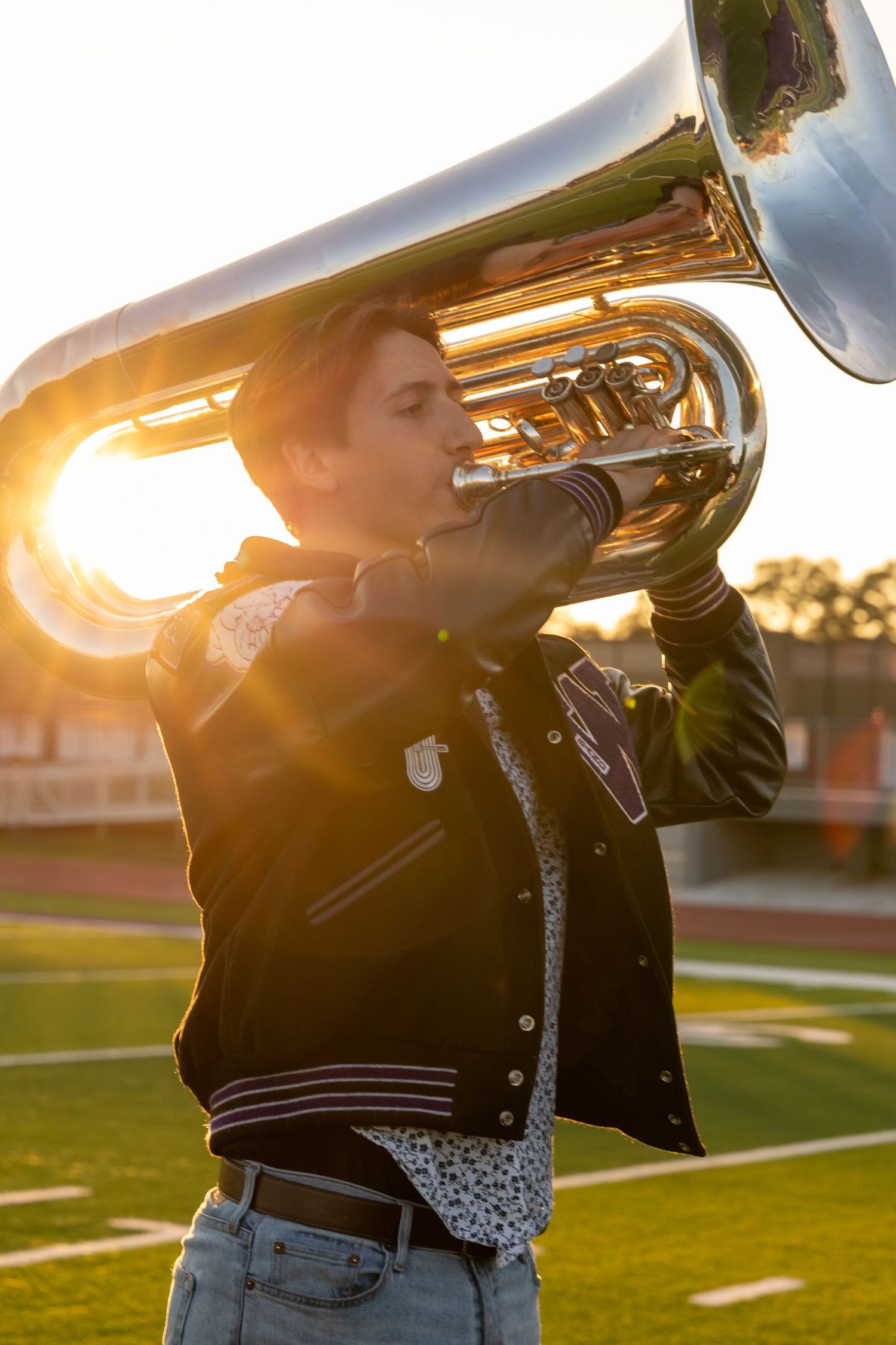 Band member plays tuba on football field during golden hour while wearing letterman jacket.