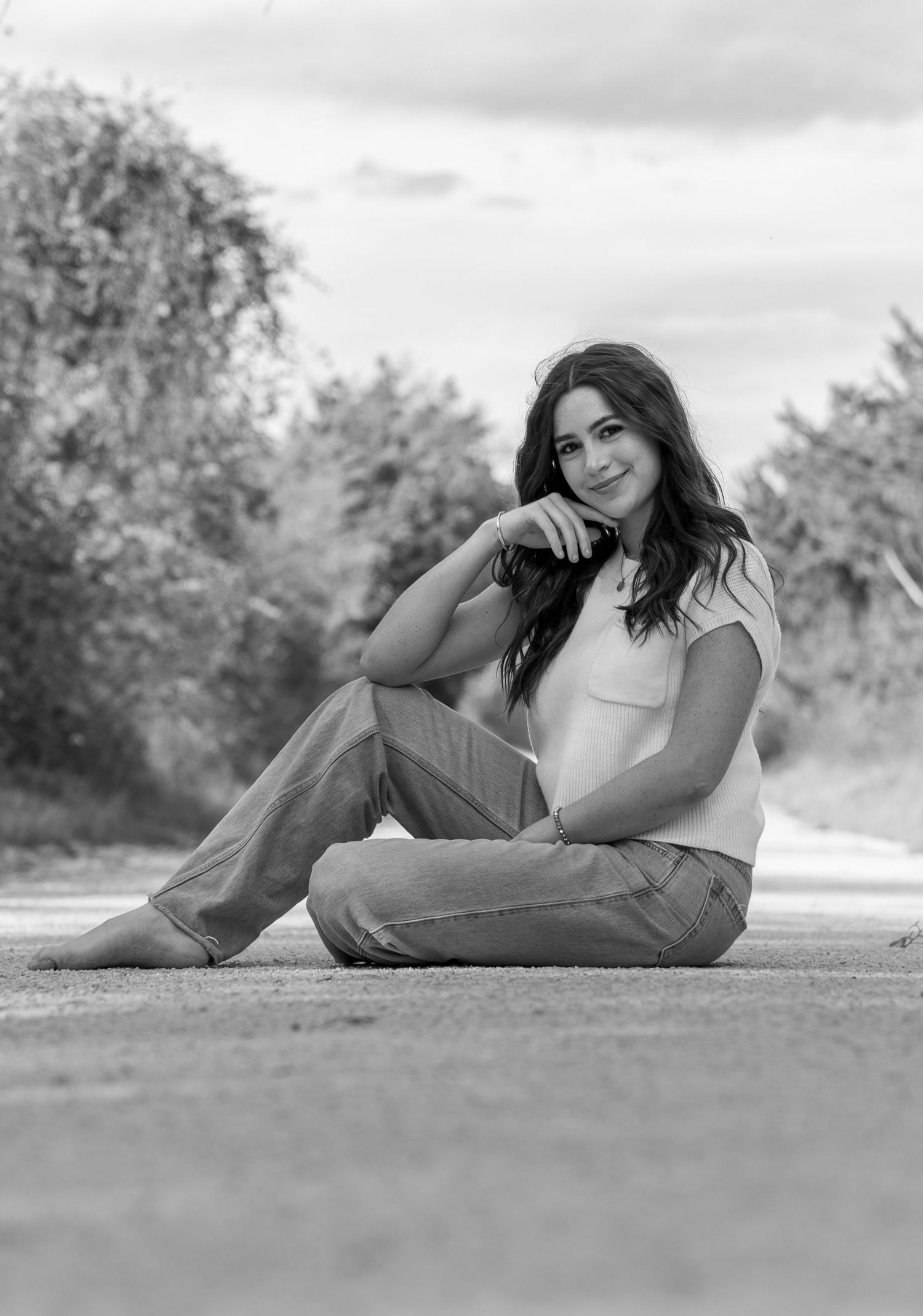 Black and white portrait of someone sitting on the ground outdoors wearing jeans and a light top in a relaxed pose.