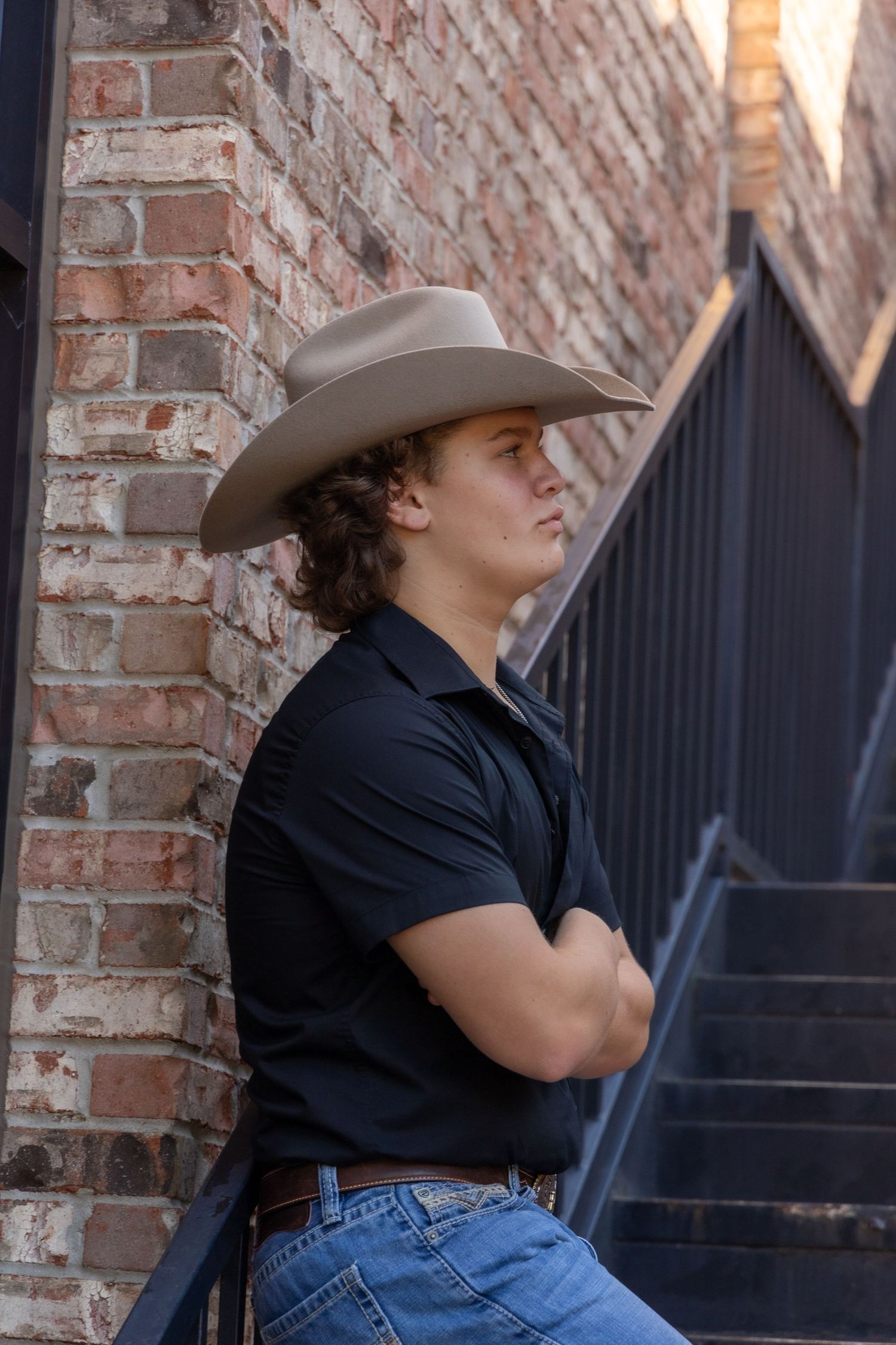 Young man with curly hair wearing cowboy hat and black shirt, leaning against brick wall with arms crossed.