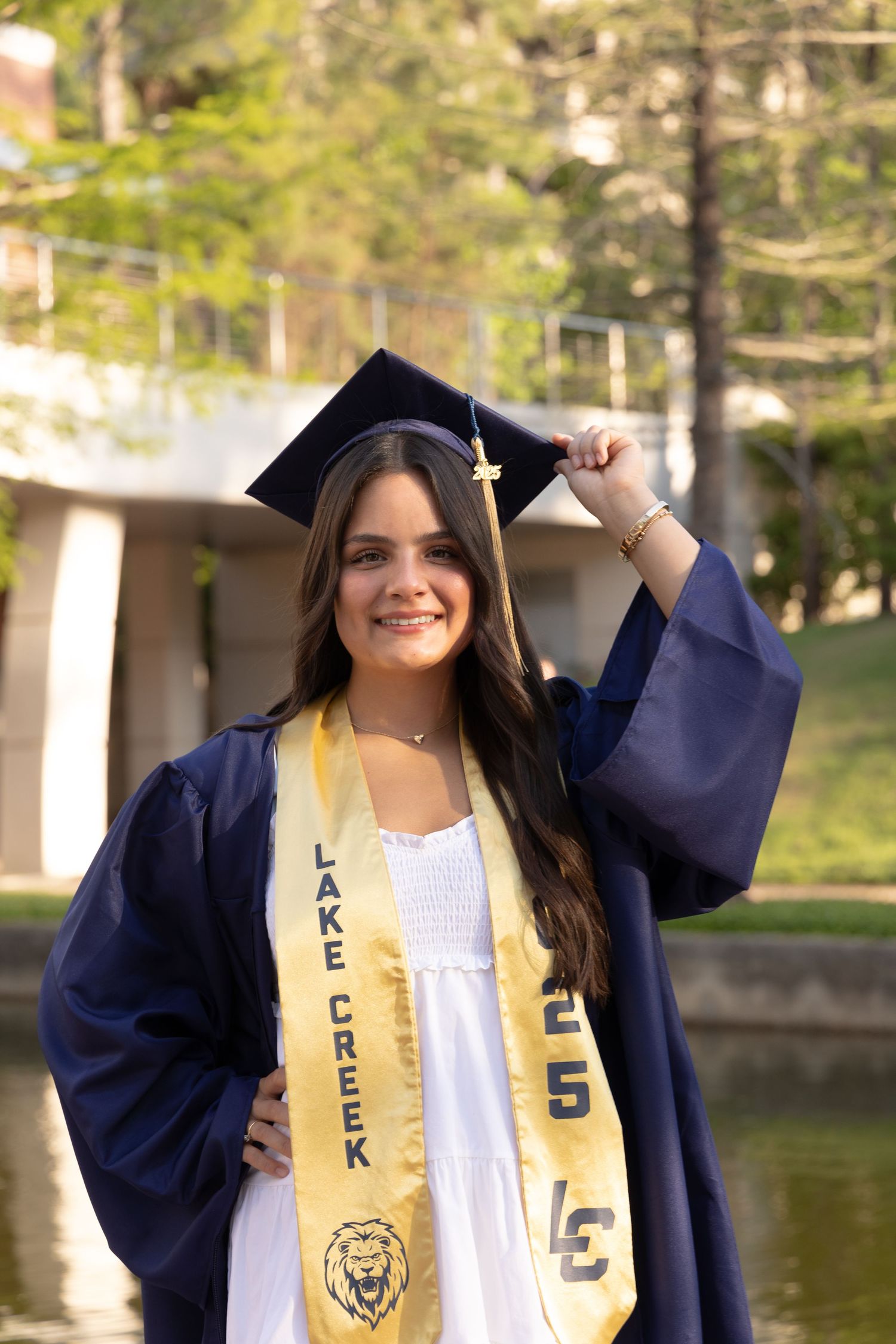 Smiling female graduate in navy cap and gown with Lake Creek Lions gold sash poses outdoors near a pond.