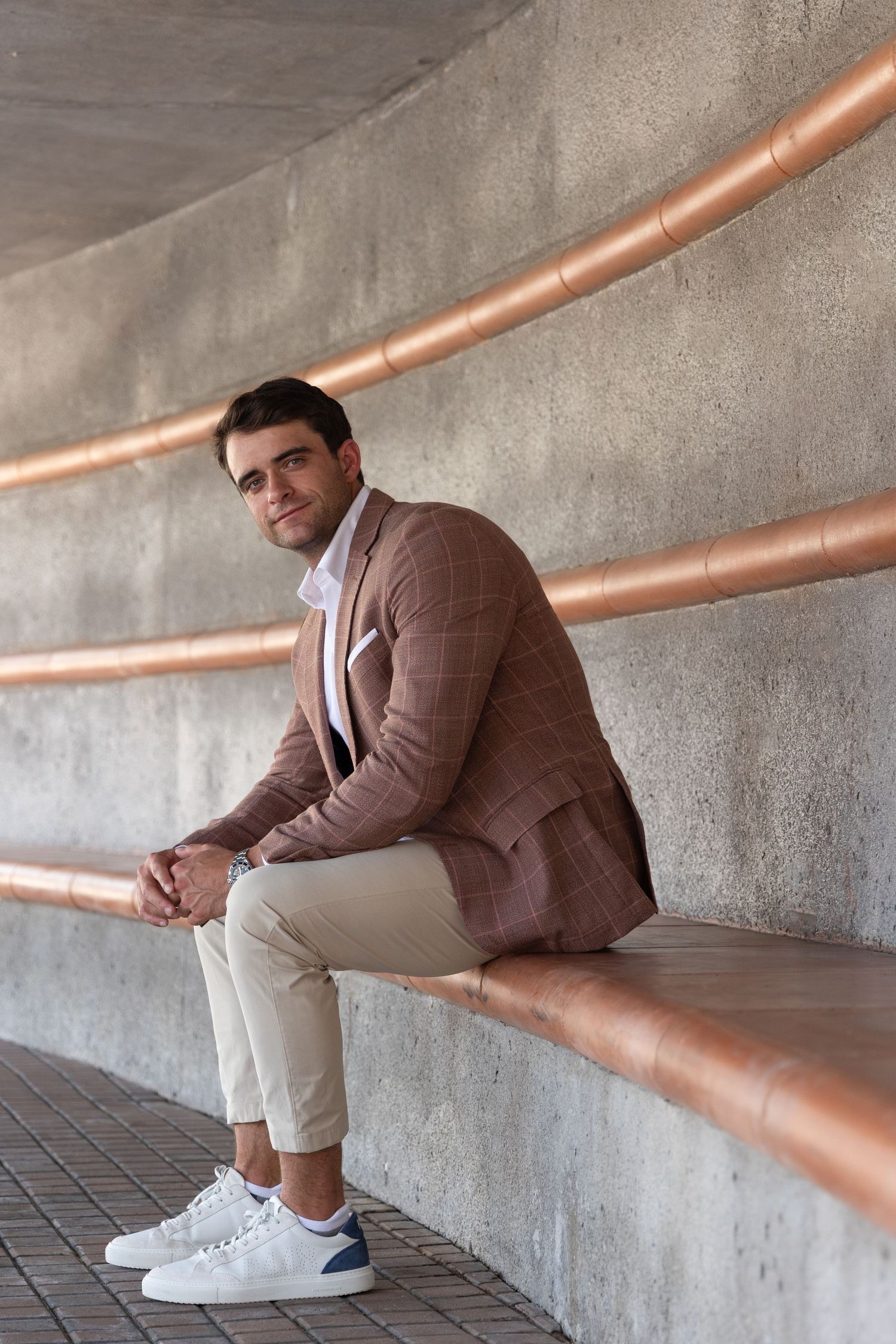 A stylish man in a plaid blazer and chinos sits on a concrete ledge with copper pipes on the wall behind him.