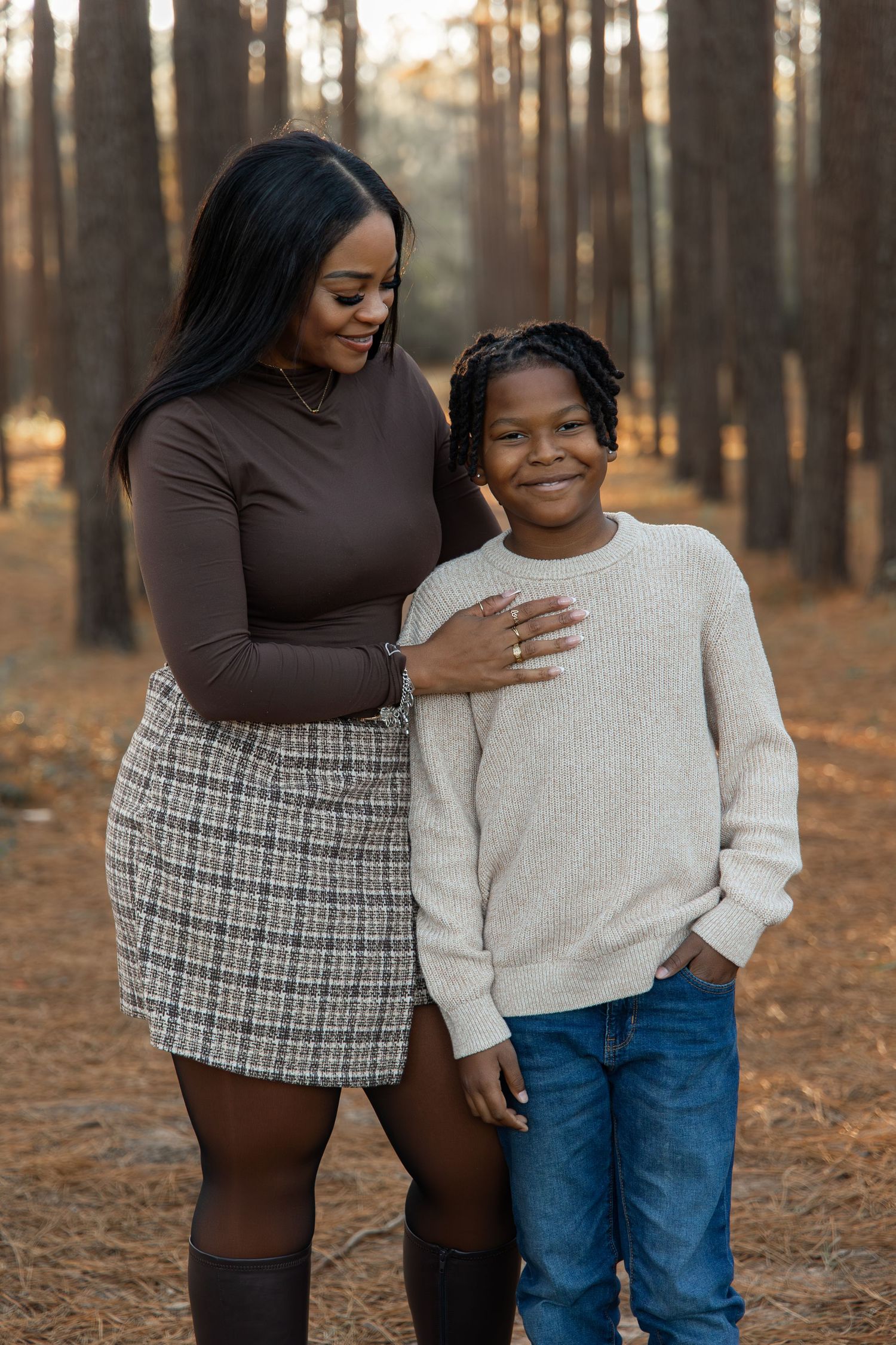 A woman and young boy pose together in a pine forest, she wears a brown top and plaid skirt, he wears a cream sweater.