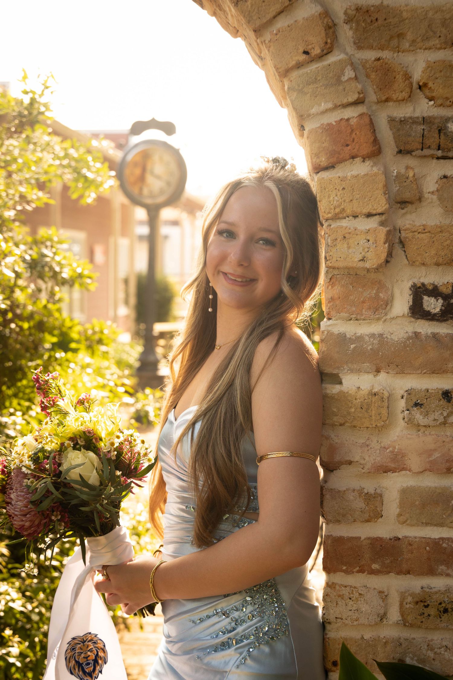 Teen girl in silver gown holding bouquet beside brick arch in golden sunlight near a street clock.