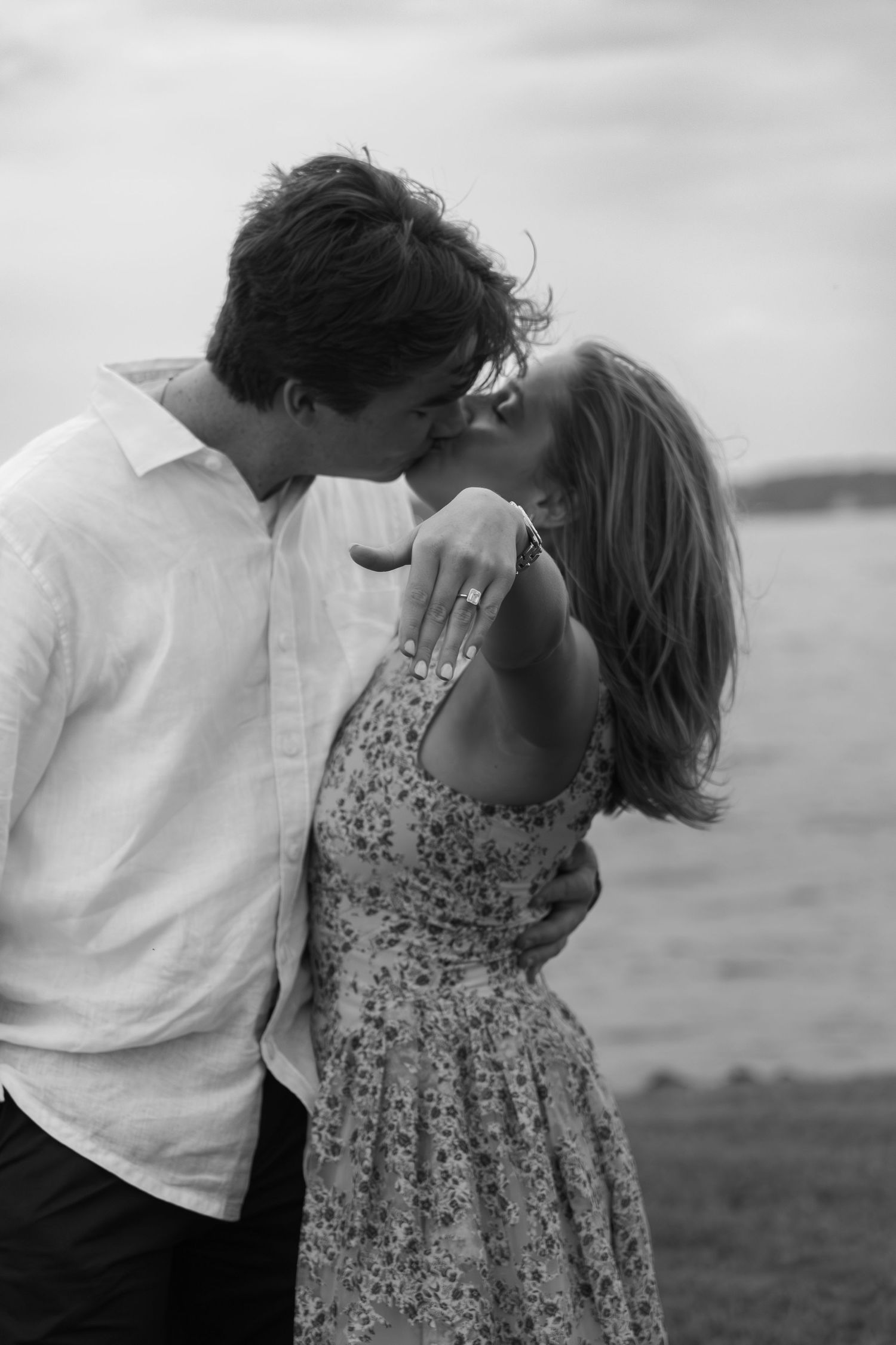 Black and white photo of a couple kissing on a beach, woman showing engagement ring, romantic seaside proposal moment.