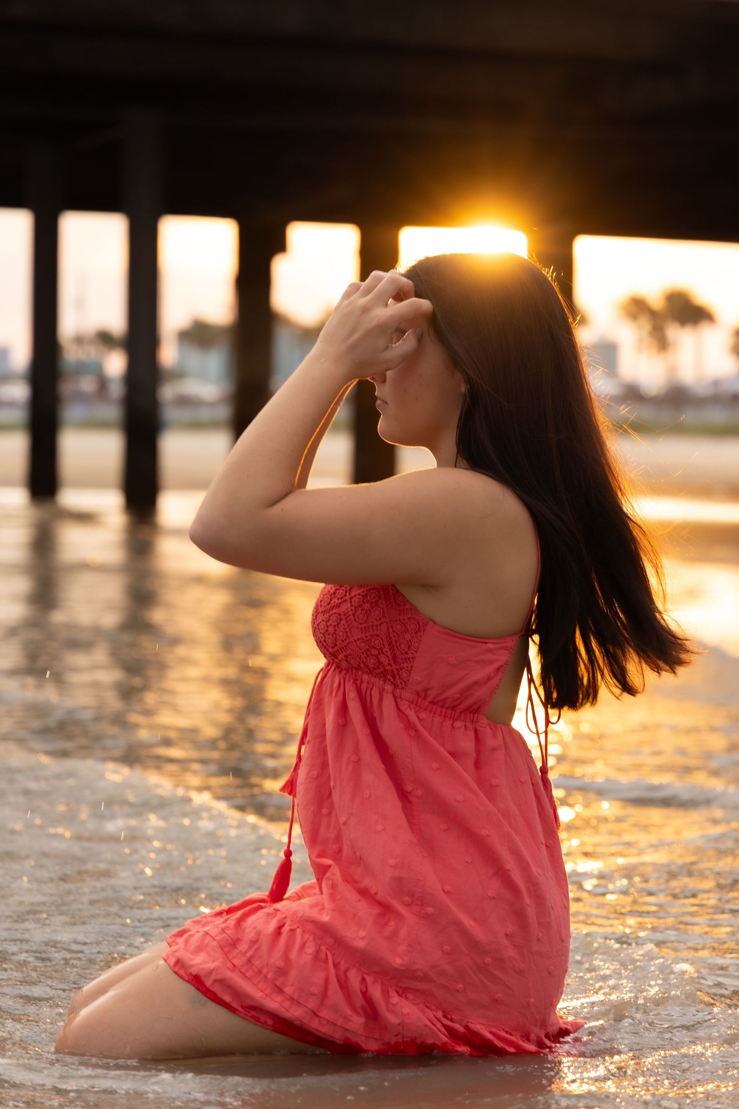 Woman in red dress kneeling in ocean water at sunset under pier with golden light glowing behind her.