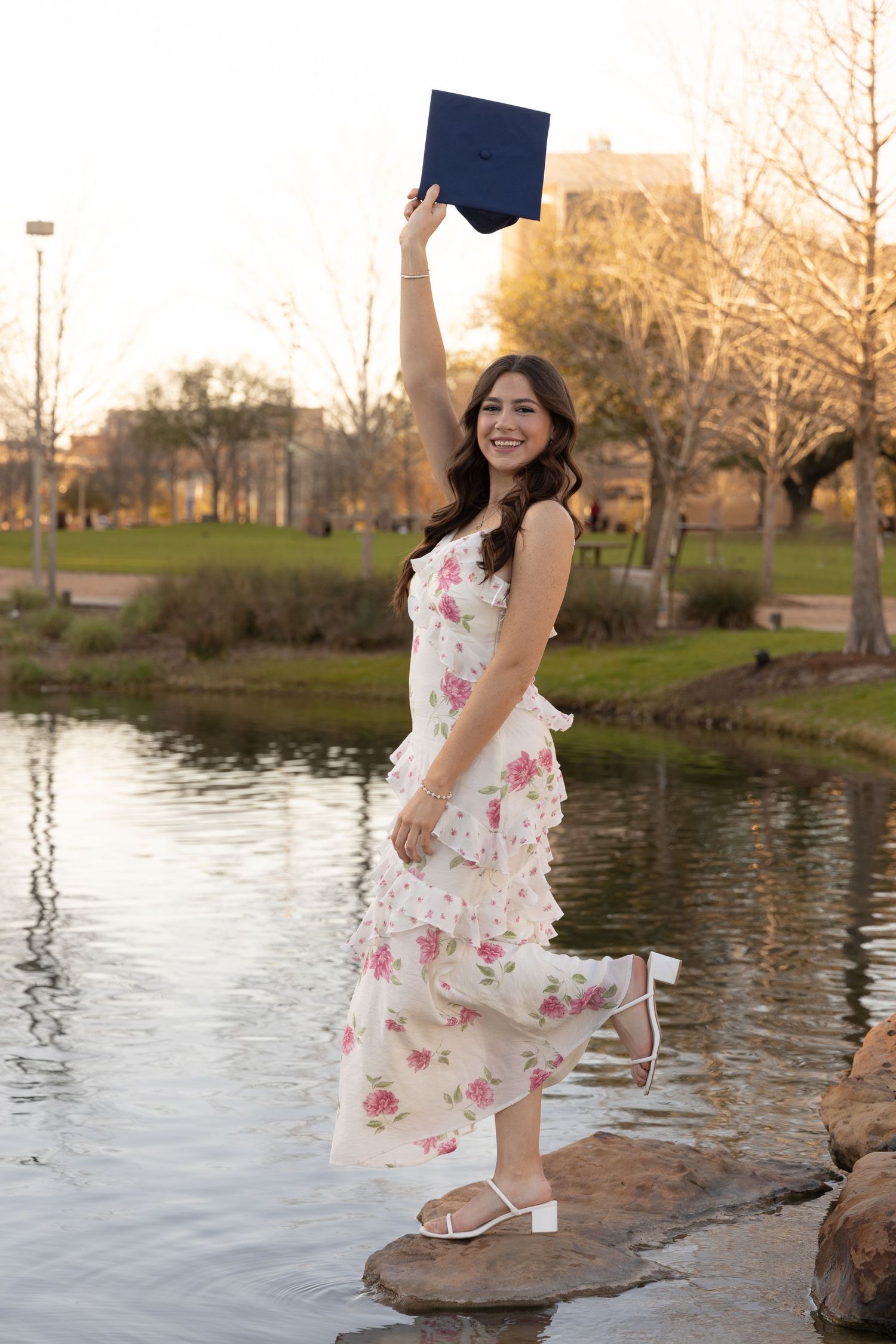 Young woman in floral dress holding graduation cap aloft, standing on rocks by a pond, celebrating her graduation outdoors.