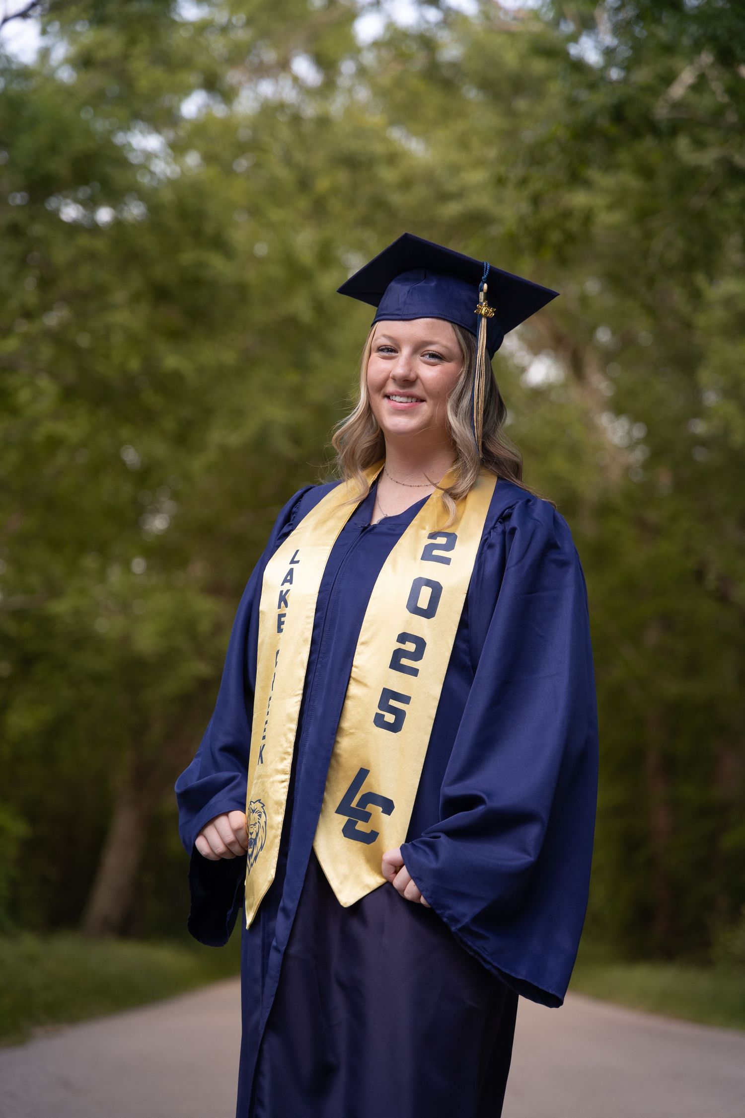 A graduate in navy blue cap and gown with gold stole poses outdoors on a tree-lined path.