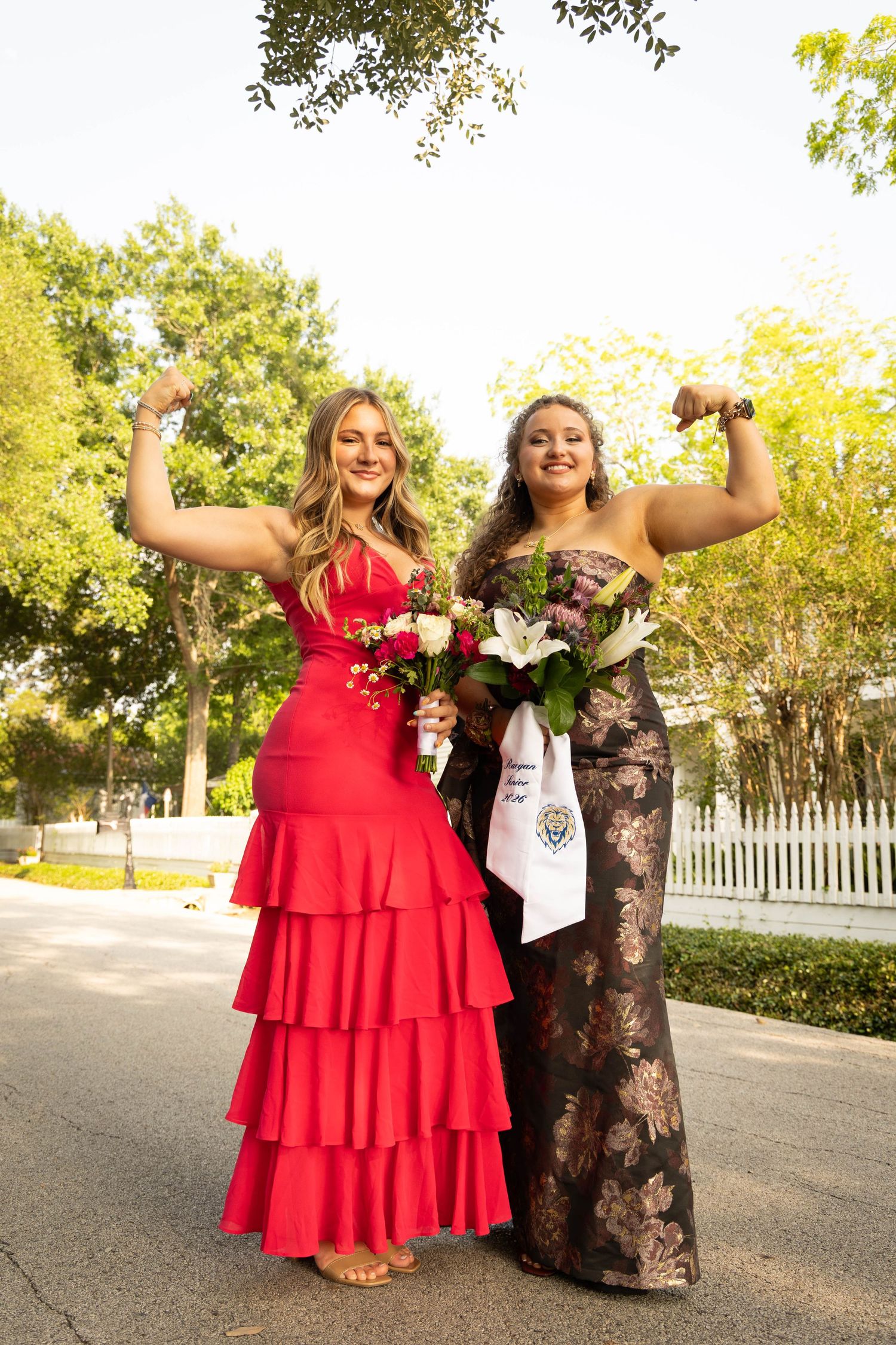 Two young women in formal prom dresses flexing muscles while holding bouquets outdoors.