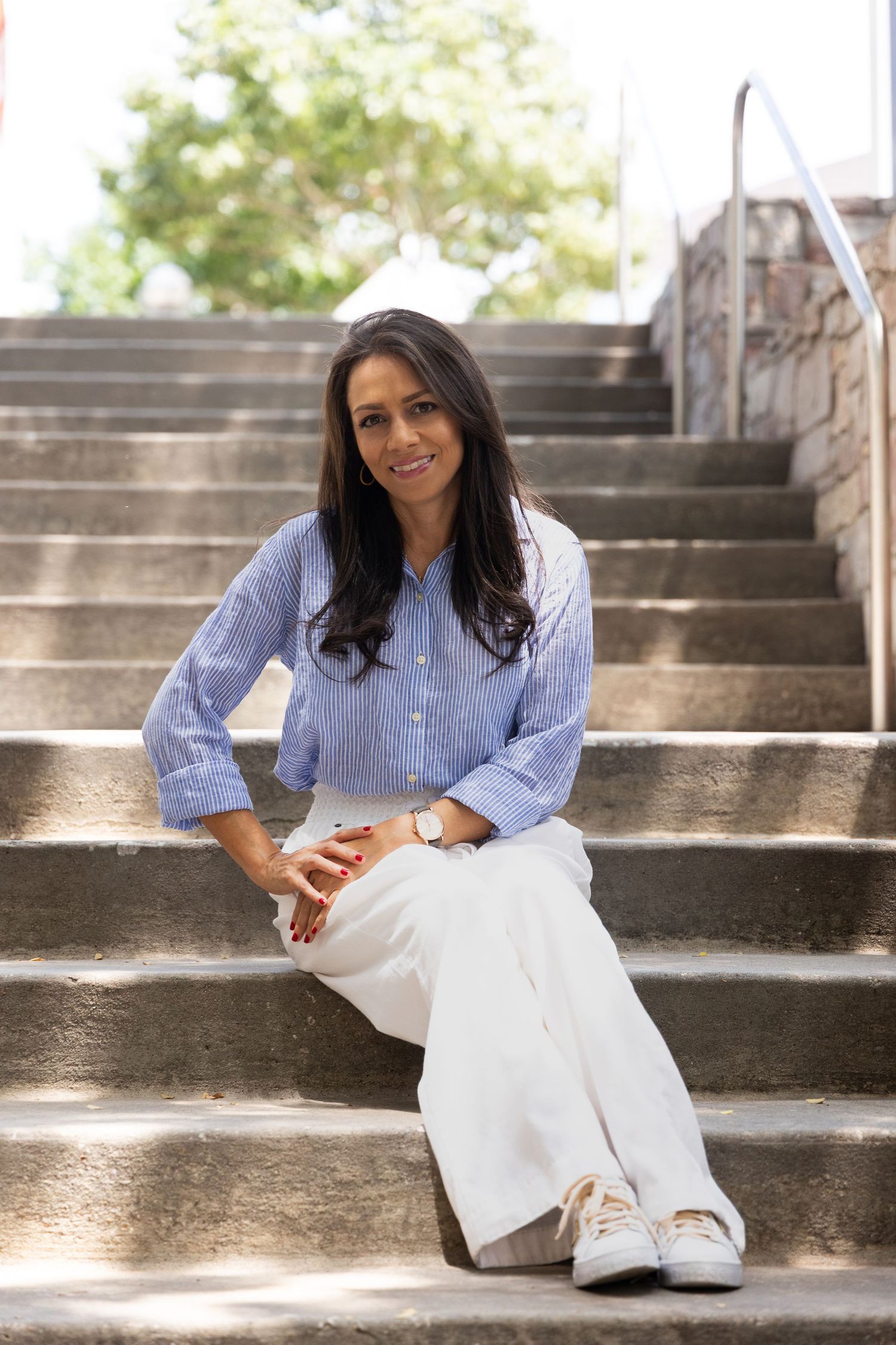 Woman in blue shirt and white pants sitting on outdoor stone steps, smiling confidently in a casual yet polished outfit.