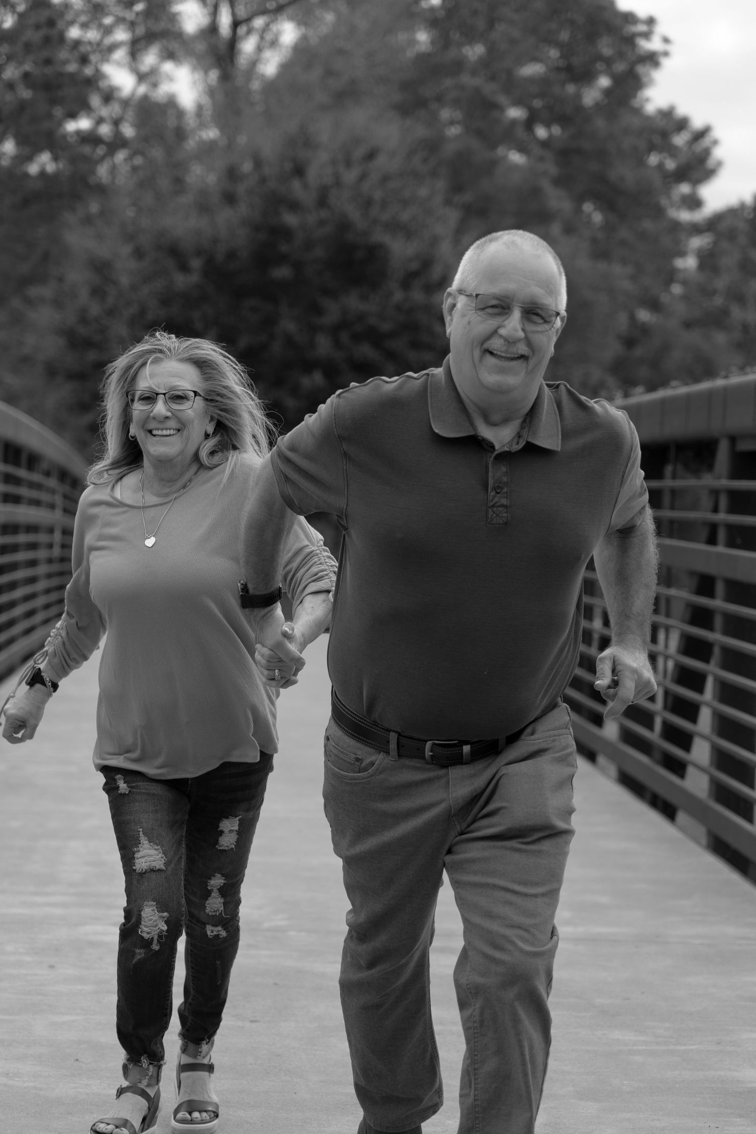 A smiling middle-aged couple holds hands while walking on a bridge outdoors, captured in black and white.