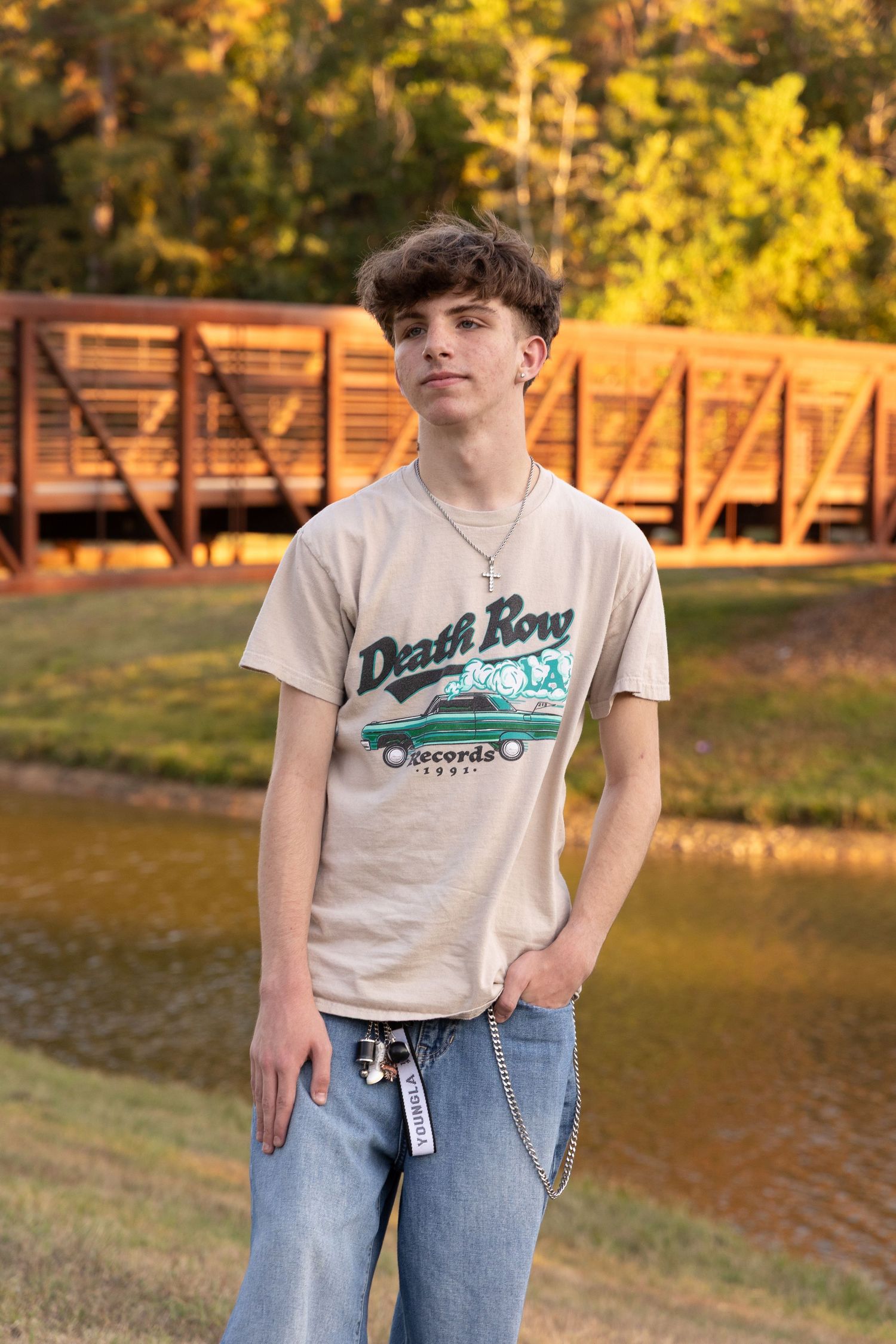 Teen in vintage Death Row Records shirt stands near bridge and pond in warm sunset lighting.