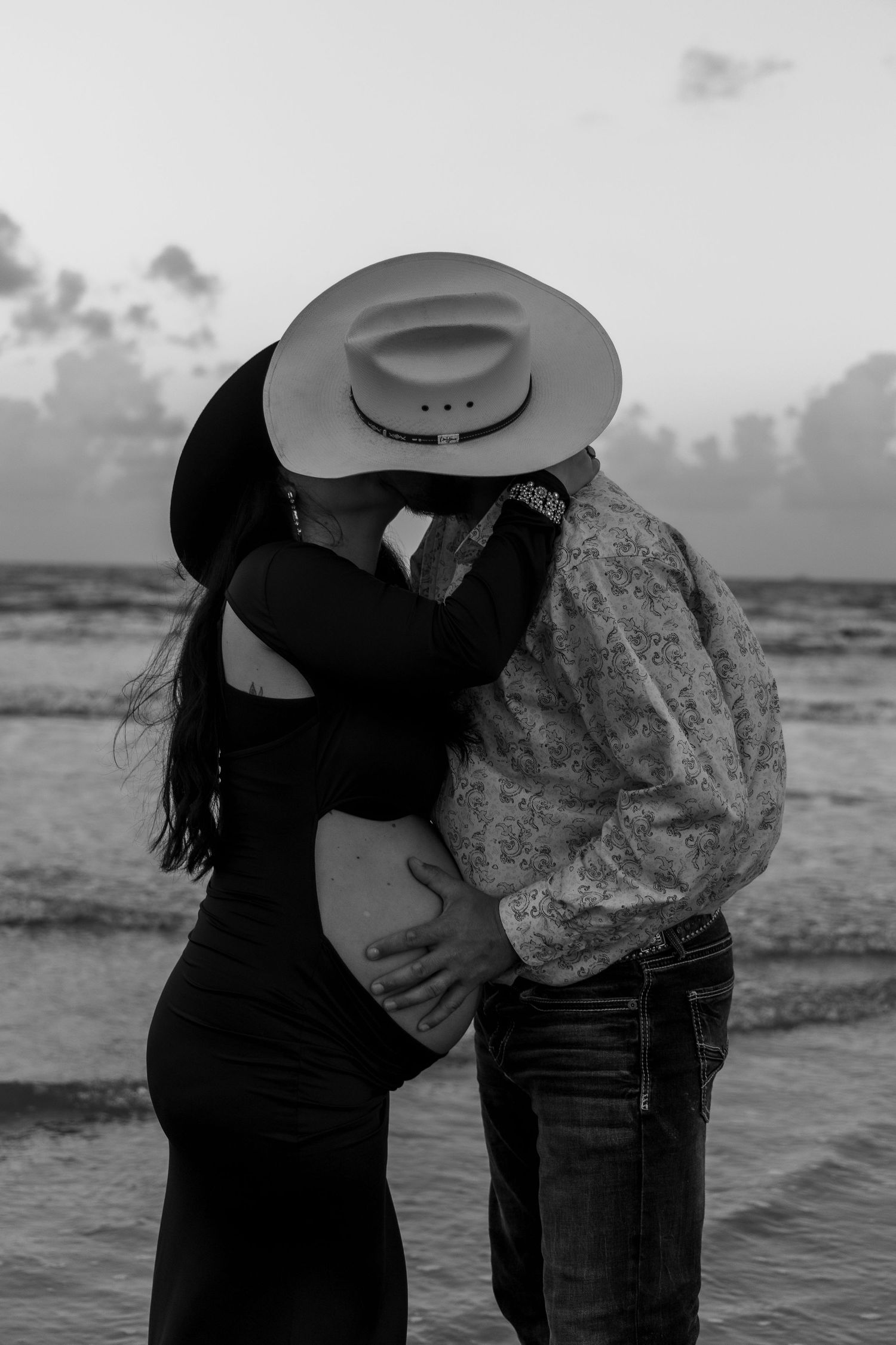 Black and white photo of a couple kissing on the beach, man in cowboy hat embracing pregnant woman in black dress.
