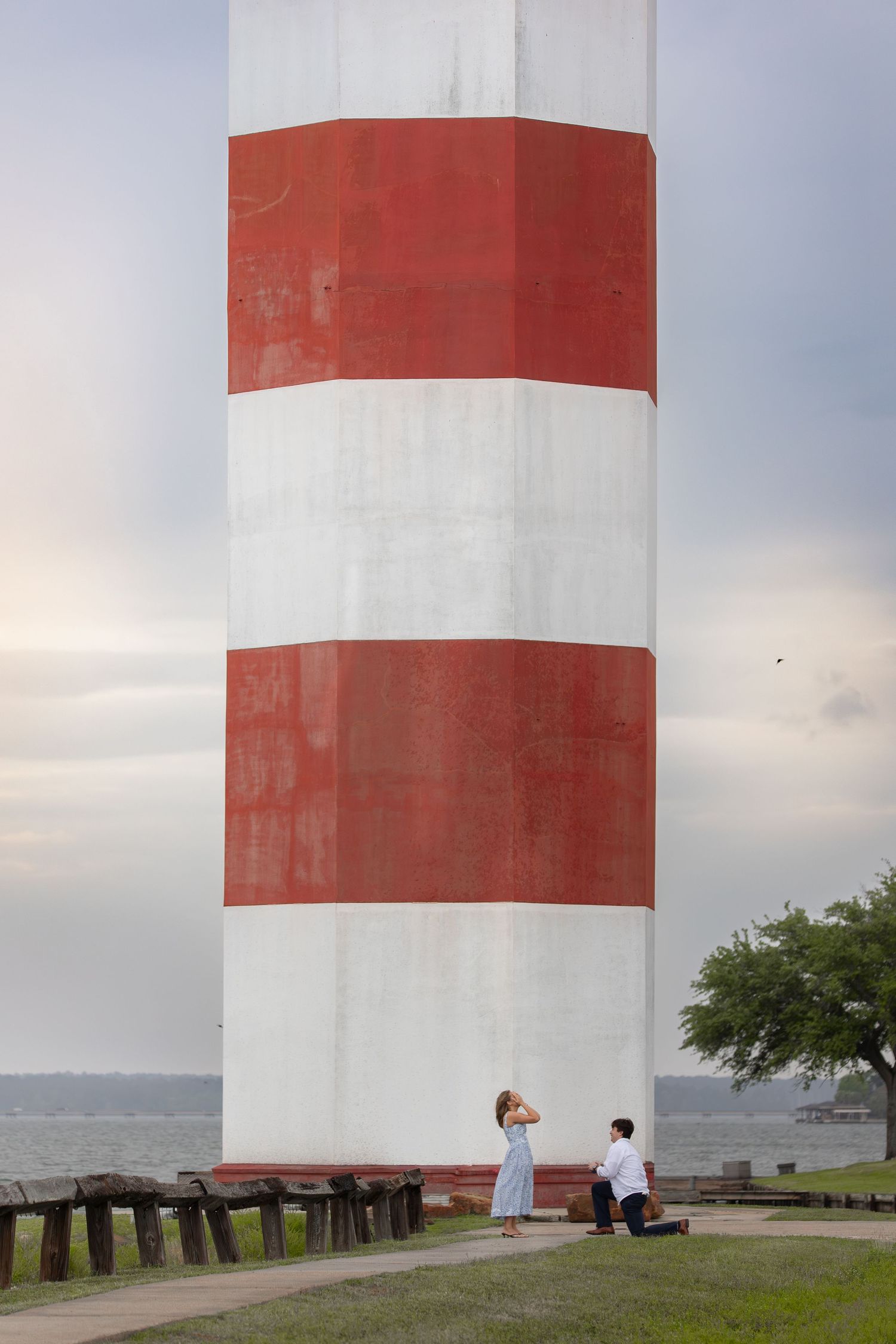 Man proposing to woman at base of red and white striped lighthouse near waterfront on cloudy day.