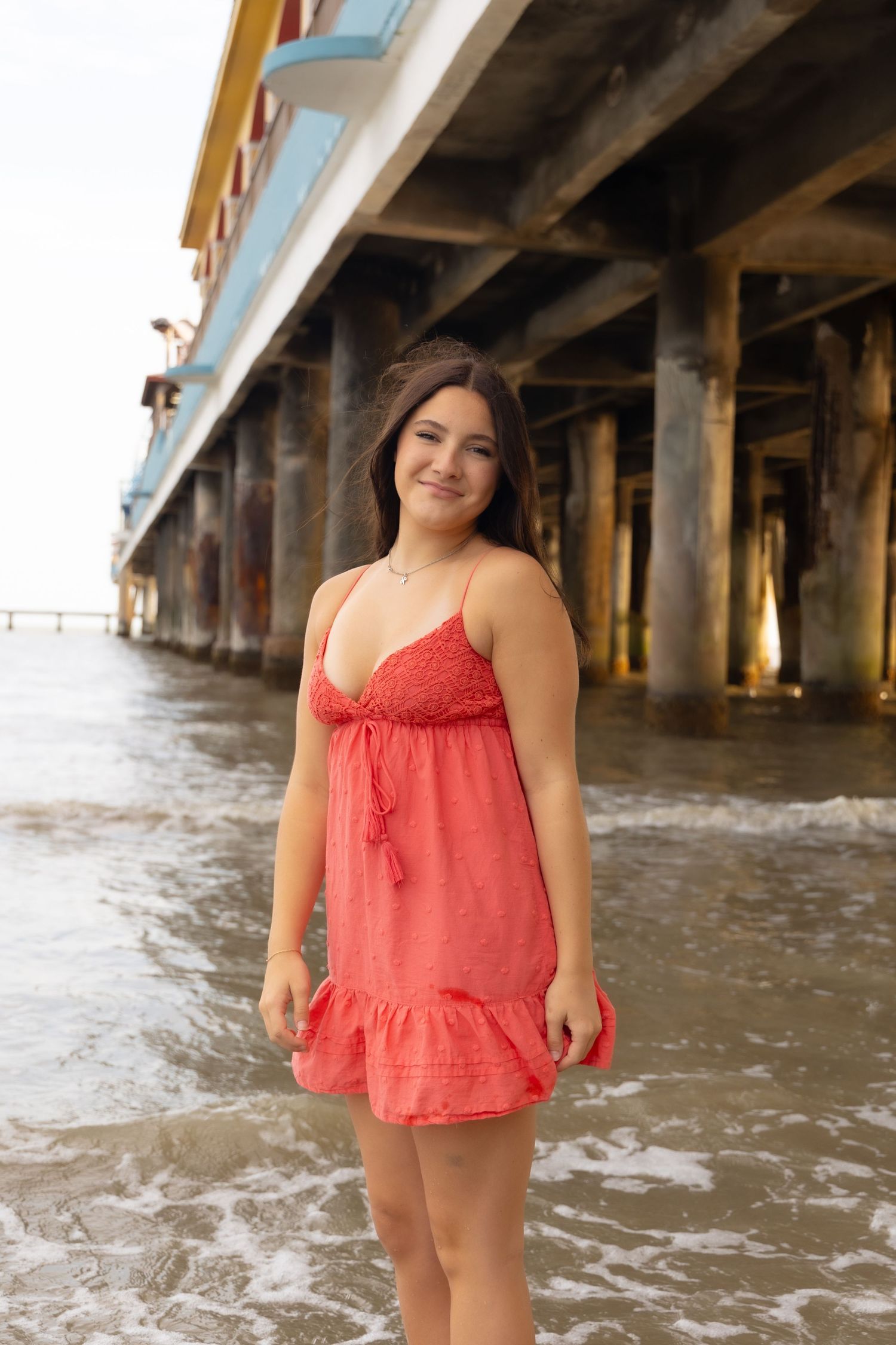 Young woman in red sundress smiling on beach near pier with ocean waves.
