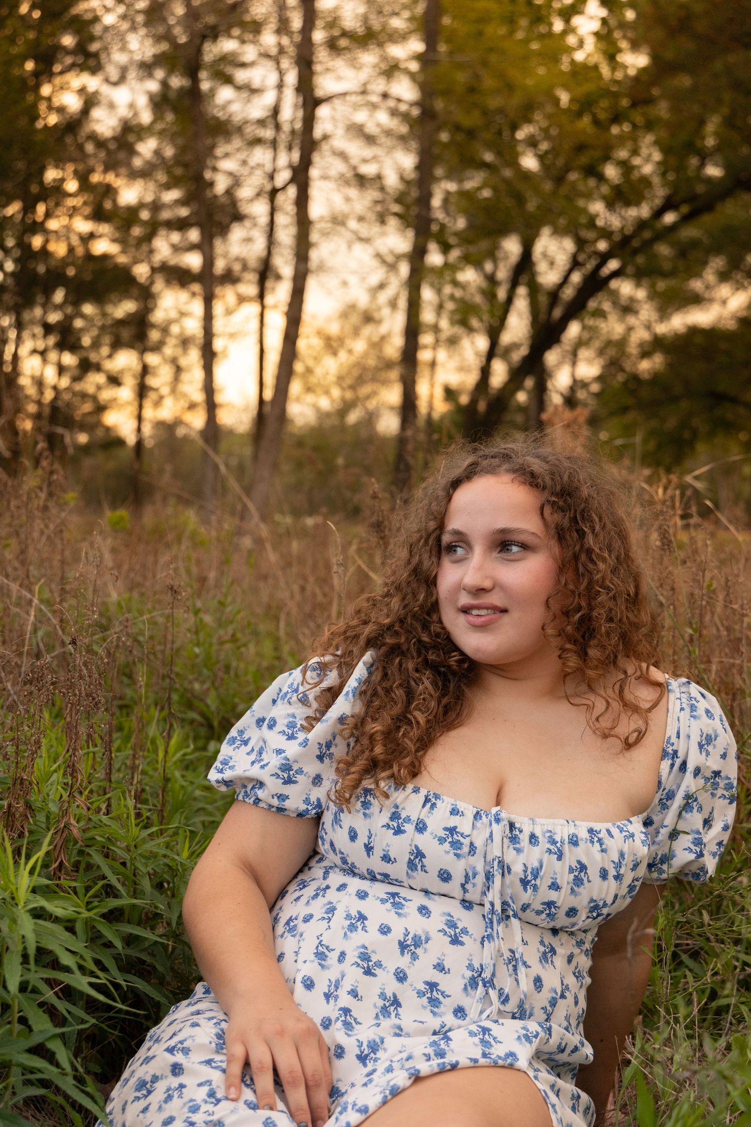 Young woman with curly hair in a floral dress sitting outdoors at golden hour in a forest meadow.
