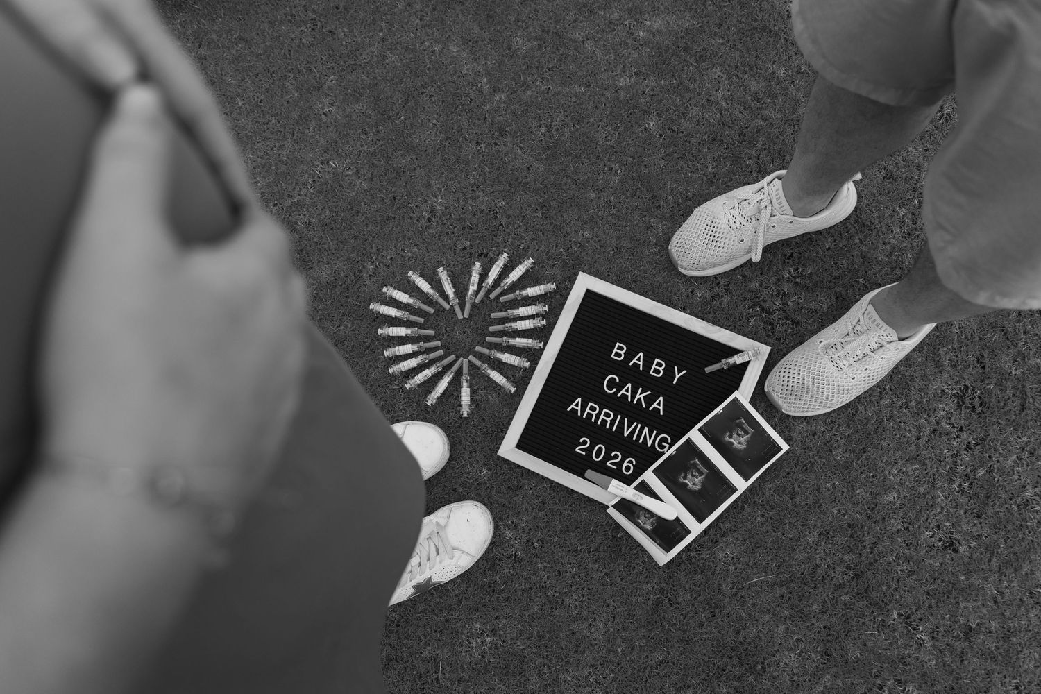 Black and white photo of couple's feet beside a letterboard sign announcing Baby Caka arriving 2026.
