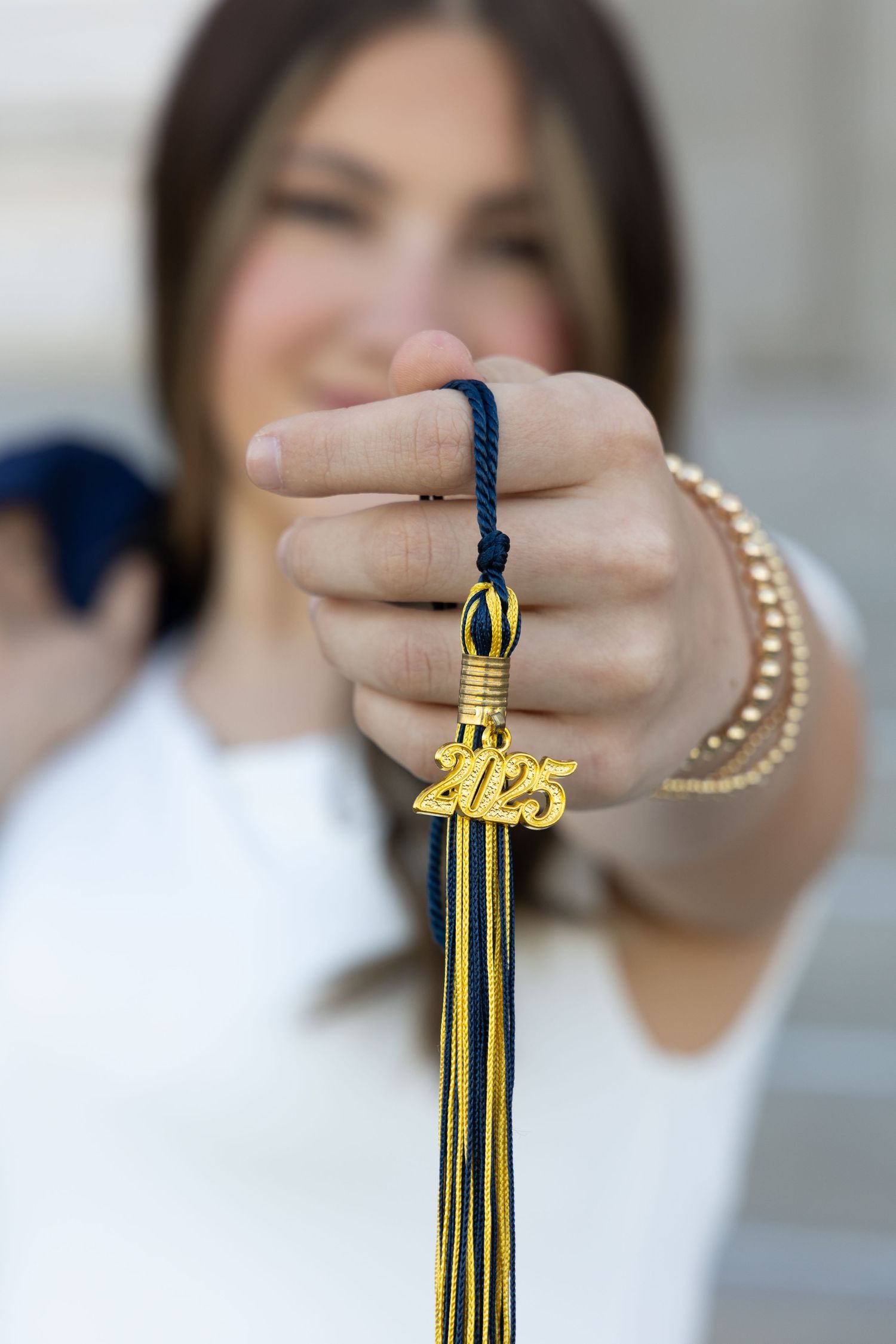 A hand holds a blue and gold graduation tassel with 2023 year charm against a blurred background.