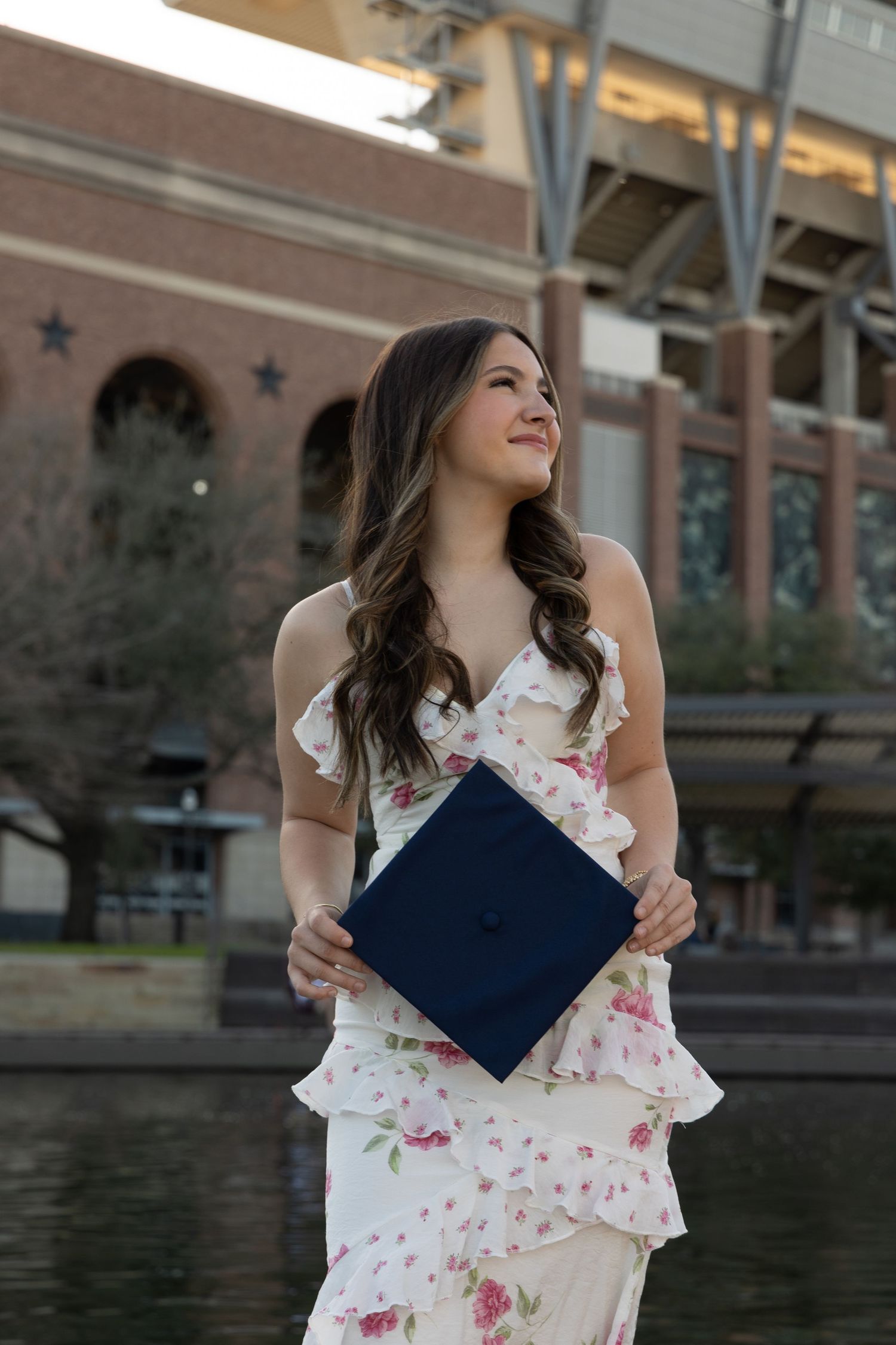 Young woman in floral dress holding navy graduation cap, smiling outdoors near brick university building.