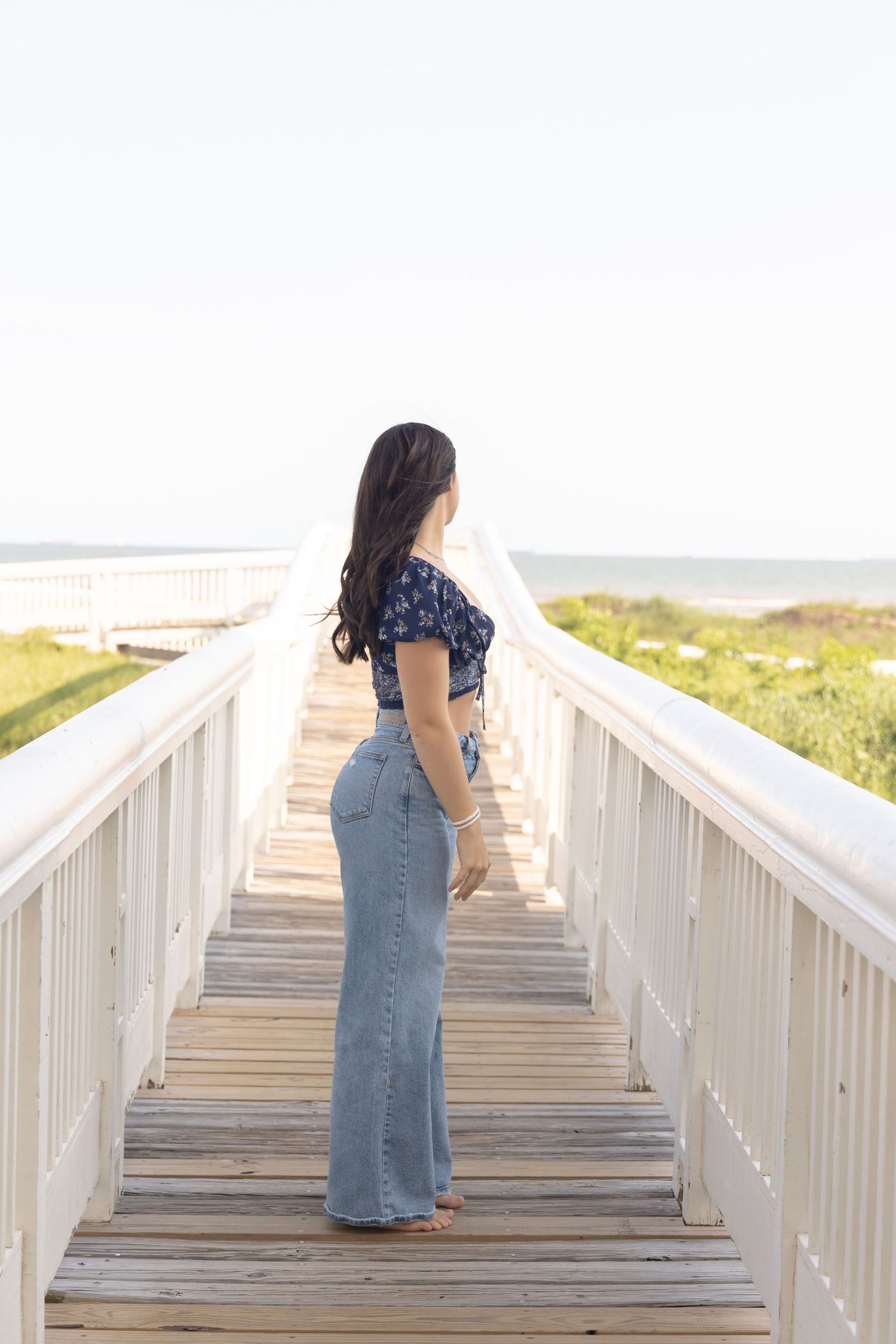 Woman in floral crop top and wide-leg jeans stands on wooden beach boardwalk, looking toward the ocean.