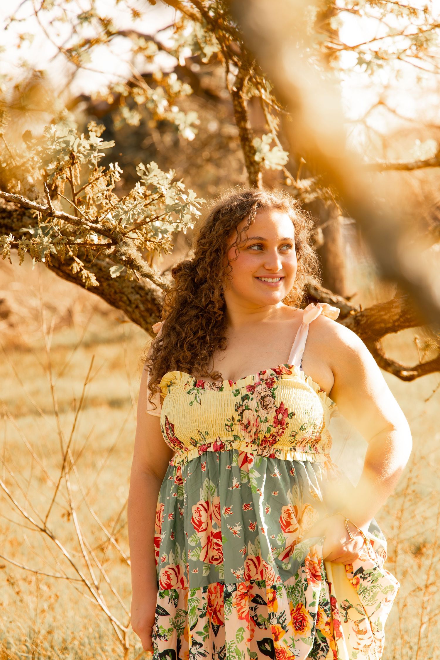 Young woman in floral dress smiling outdoors among golden trees in warm sunlight.