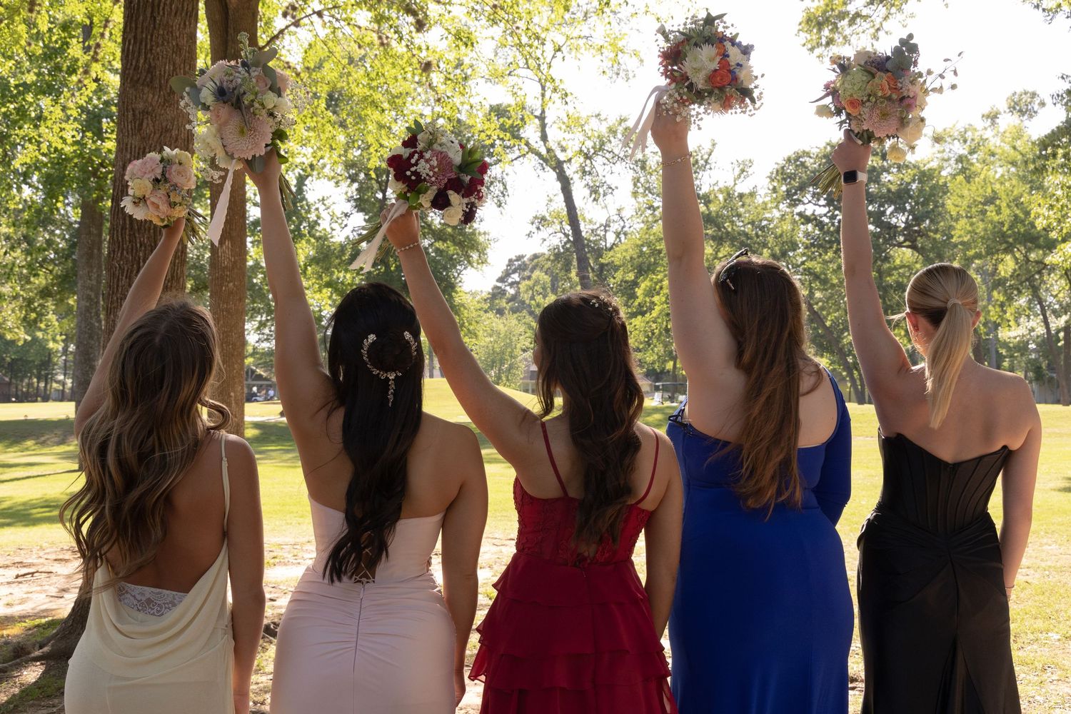 Five bridesmaids in colorful dresses raise bouquets outdoors in a sunny park setting.