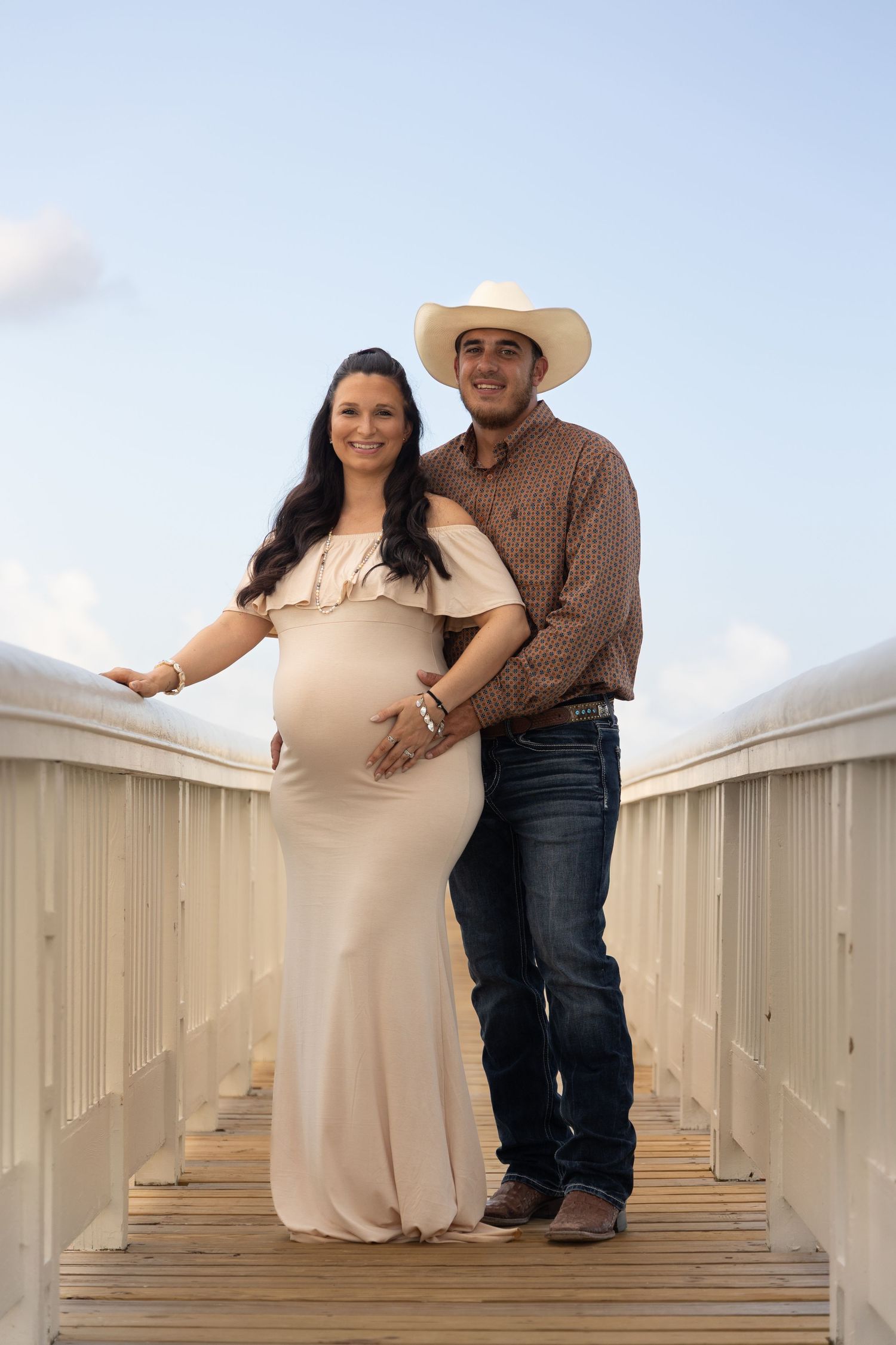 Pregnant woman in beige off-shoulder gown with man in cowboy hat on wooden bridge at sunset.