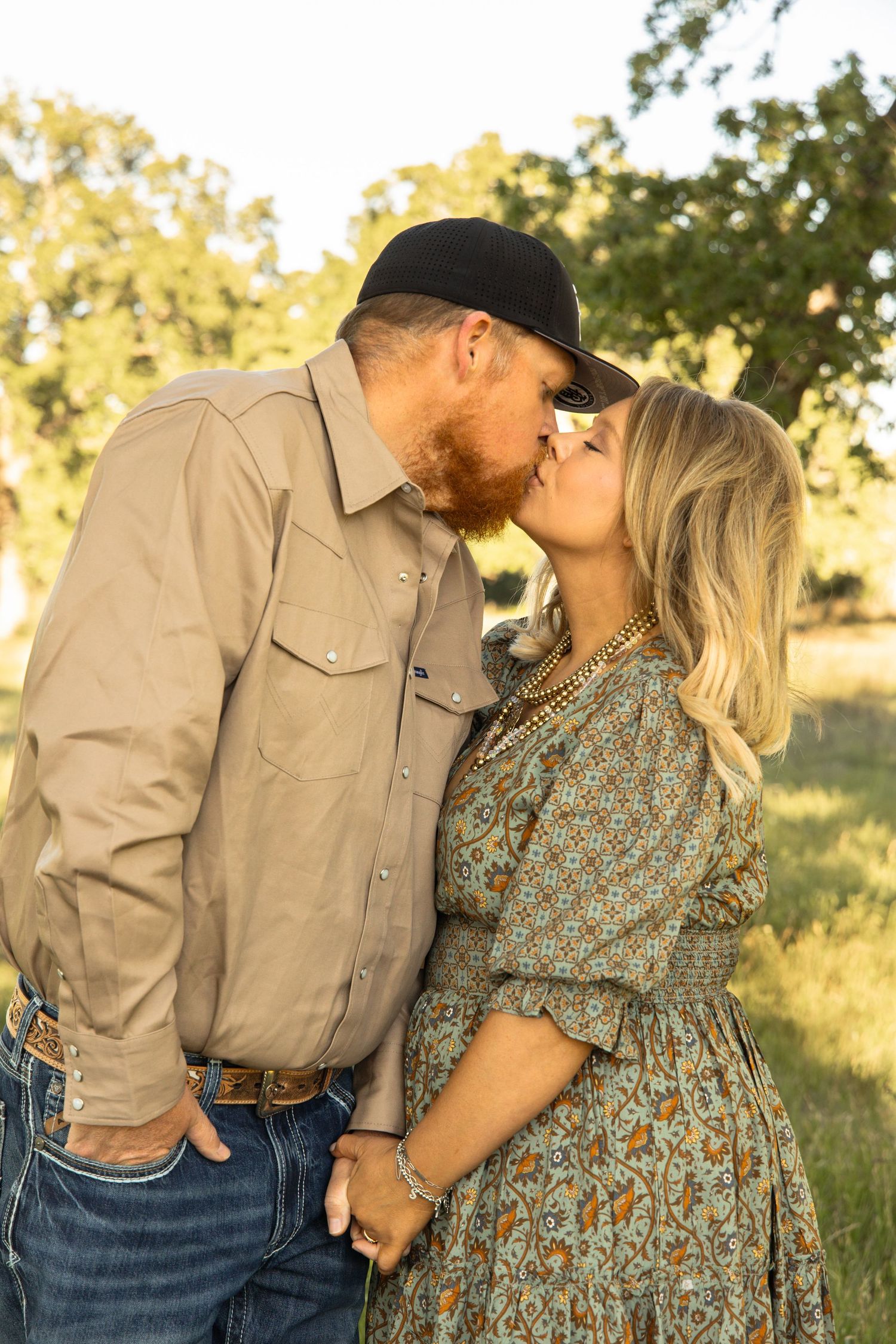 A couple sharing a romantic kiss outdoors, man in tan shirt and black hat, woman in floral dress, green trees in background.